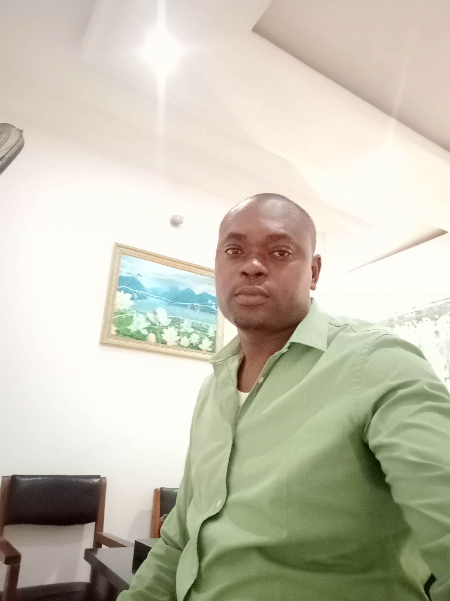 A medium shot captures a man in a green collared shirt, positioned indoors against a white wall. A framed painting hangs on the wall behind him. Two dark wooden chairs are visible in the lower left corner. The setting appears to be indoors, possibly a home or office, but specific details to confirm the location in Gwarinpa, Nigeria, are absent. No actions or interactions are discernible from the single image.