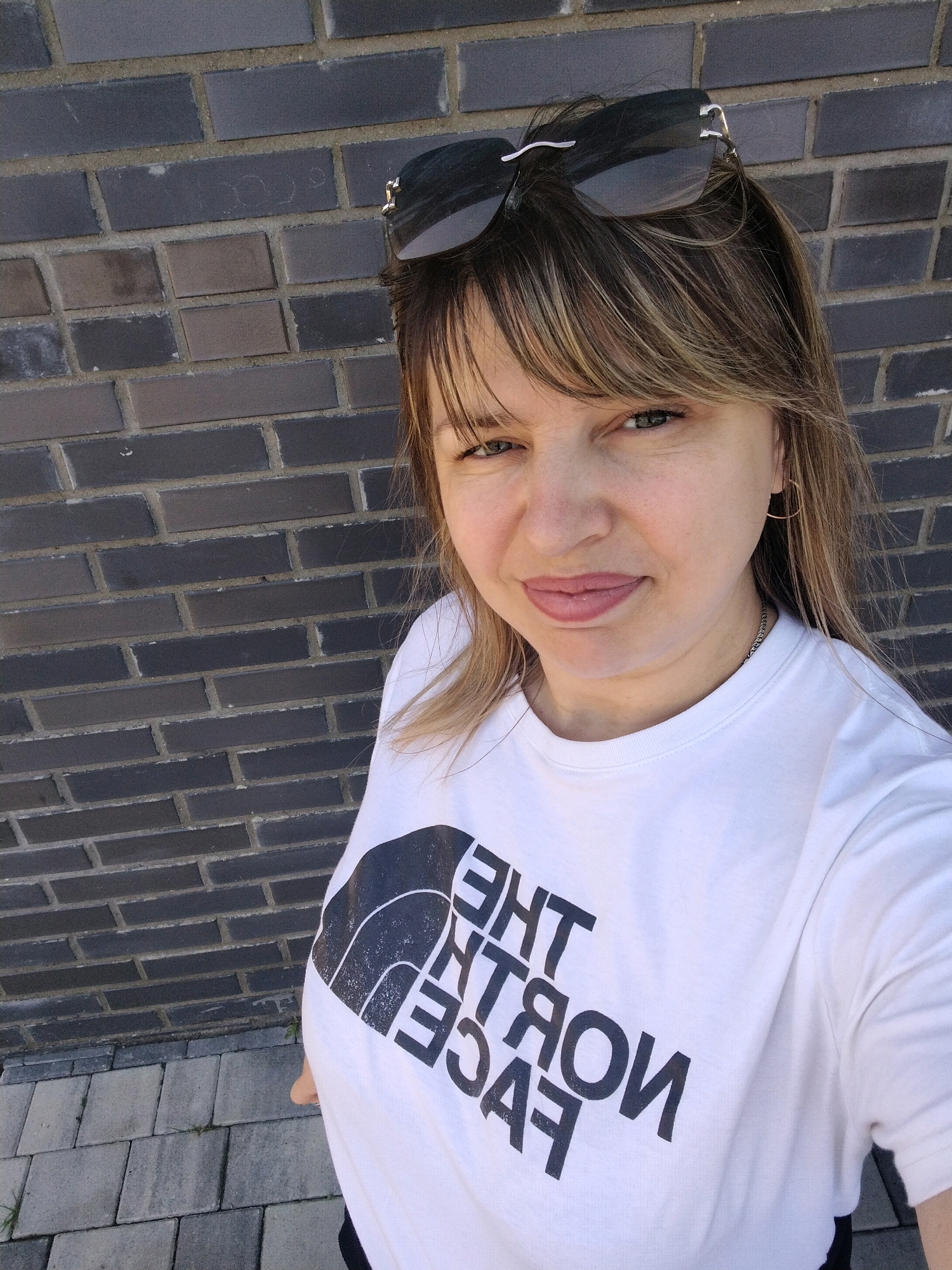 The image is a close-up selfie of a woman with blonde hair, wearing sunglasses on her head and a white t-shirt with "The North Face" logo printed on it. She is standing in front of a brick wall. The lower part of the image shows a paved walkway. The image does not contain any information that could place it in Yaroslavl, Russia.