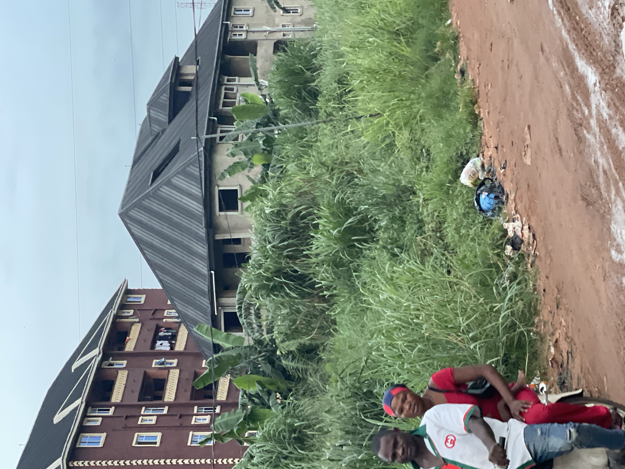 The image, taken in Nkpor, Nigeria, shows two buildings, one light gray with a multi-angled roof and another brown with multiple rectangular windows, adjacent to a patch of overgrown green vegetation. The vegetation encroaches onto a dirt road with light patches. Discarded waste is visible along the roadside. Two dark-skinned individuals are positioned in the foreground. One is dressed in a white shirt with green and red accents and denim jeans. The second individual is wearing a red outfit and a blue head covering. Both individuals appear to be seated on a parked motorcycle.