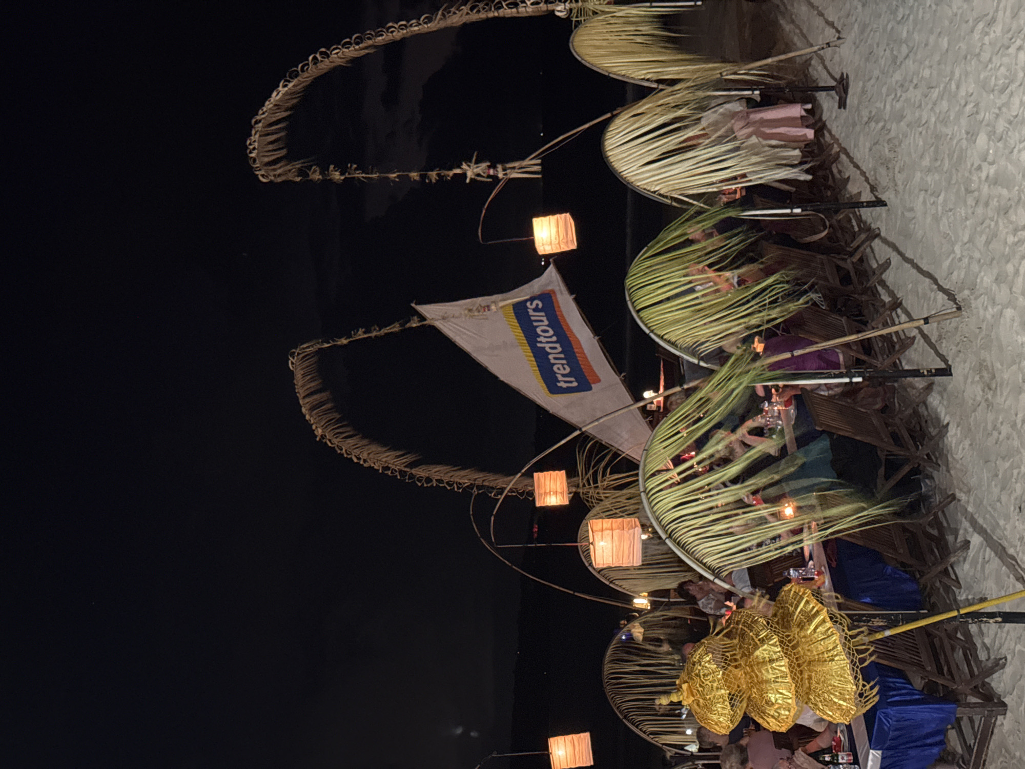 The image depicts an outdoor dining scene at night on a sandy beach, likely in Kuta Selatan, Indonesia, given the tropical decor. Several tables are set for dinner, covered with blue tablecloths and adorned with white placemats and silverware. Wooden chairs are arranged around the tables. The setting is illuminated by square, orange paper lanterns hanging from bamboo structures and palm fronds that arch over the dining area, creating a natural canopy. In the background, a white flag with the blue and orange logo "trendtours" is visible, supported by bamboo poles. People are seated at the tables, engaged in dining and conversation. A prominent gold, multi-tiered decorative umbrella-like object stands to the left of the frame. The overall atmosphere suggests a festive or celebratory beachfront dining experience.