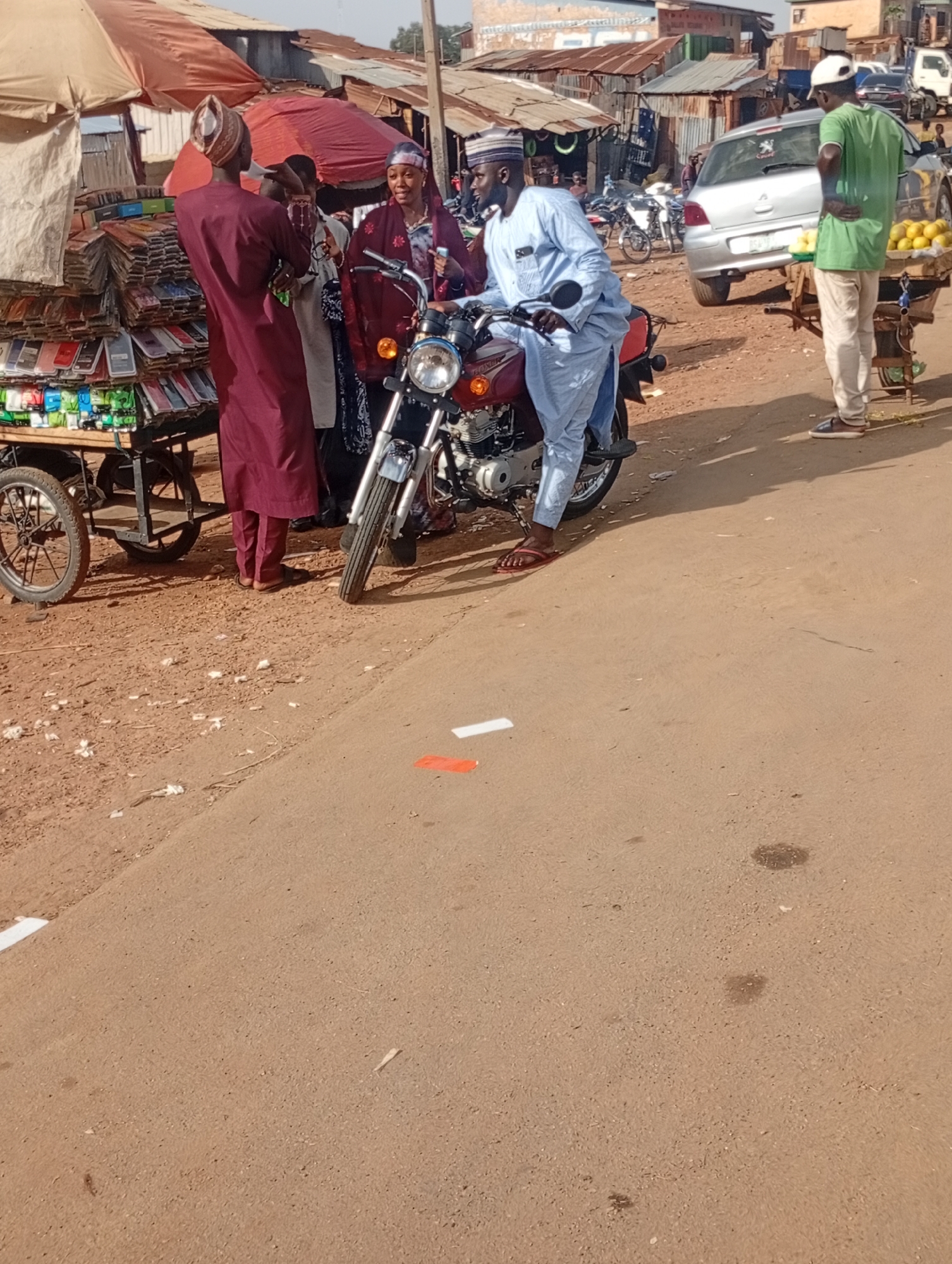 In the city of Rafin Par, Nigeria, on a dirt road, a group of individuals are gathered around a motorcycle. A man in a light blue robe and a white cap is seated on the motorcycle, holding onto its handlebars. To his left, a man in a maroon robe and a patterned cap is talking to him. A woman in a patterned red garment and a headscarf stands behind them, holding something in her hand and looking at the man on the motorcycle. In the background, a cart laden with goods, possibly phone accessories, is visible, along with more people and market stalls. Further back, a silver car and a fruit stand with yellow fruits on display are present, as well as more market structures and vehicles. The environment appears to be a bustling open-air market with a mix of permanent and temporary structures. The ground is composed of dirt and some scattered debris. White and orange markings are visible on the road surface.
