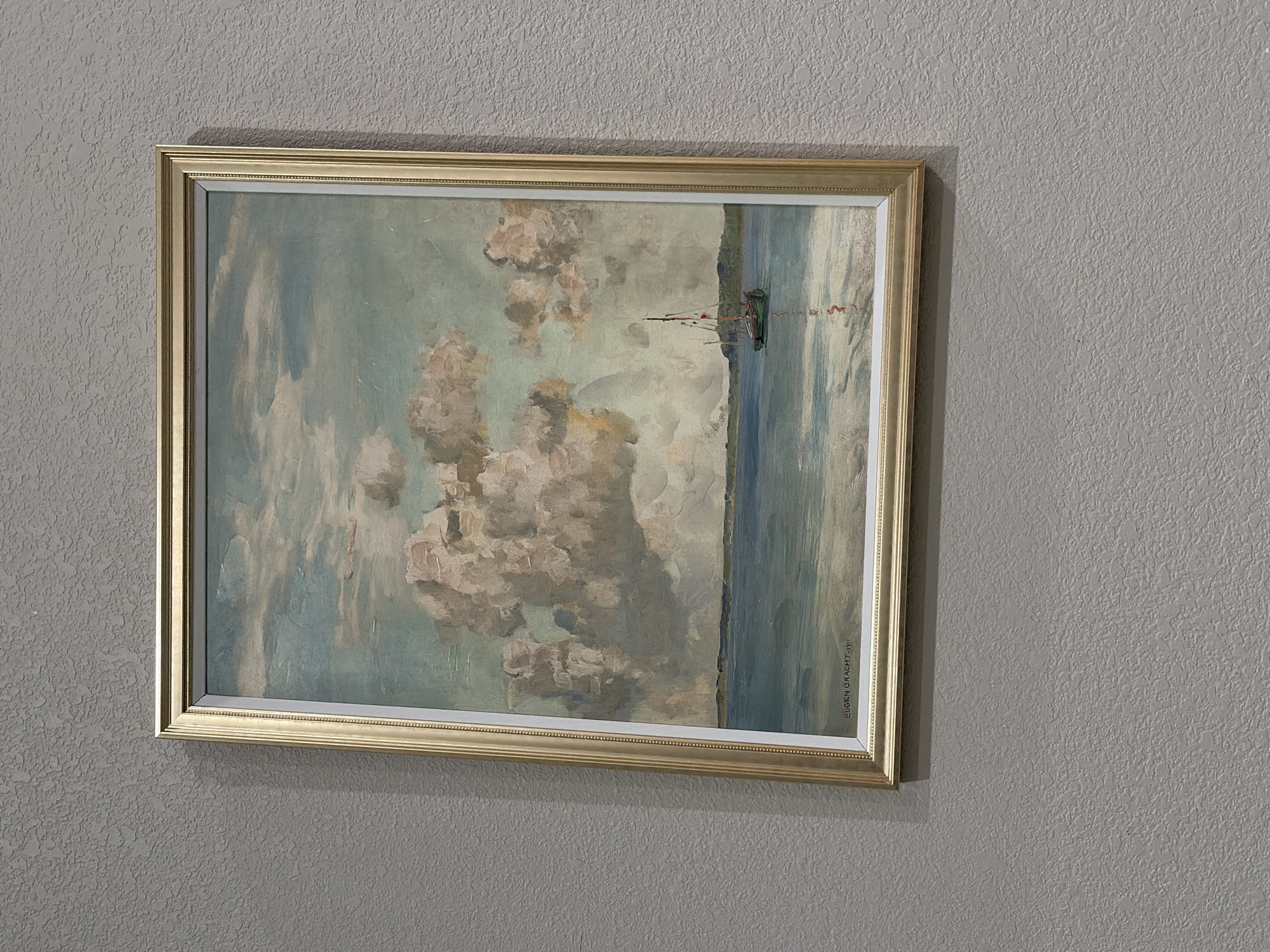 A framed painting hangs on a textured grey wall. The painting depicts a serene landscape with a body of water on the right and a cloudy sky on the left. The water is rendered in shades of blue and grey, with vertical brushstrokes suggesting movement. On the far right edge of the painting, a dark green object with red accents appears to be a small boat or buoy in the water. The sky is filled with large, billowy clouds in tones of white, grey, and pale yellow, with hints of blue peeking through. The overall impression is one of a calm, possibly overcast day. In the bottom right corner of the painting, a signature is visible: "EUGEN BRACHT 1911". The frame is a simple gold-colored molding with a subtle patterned inner edge. The textured wall creates a backdrop that emphasizes the artwork. The lighting suggests it could be daytime, with soft light illuminating the painting and casting a gentle shadow on the wall.