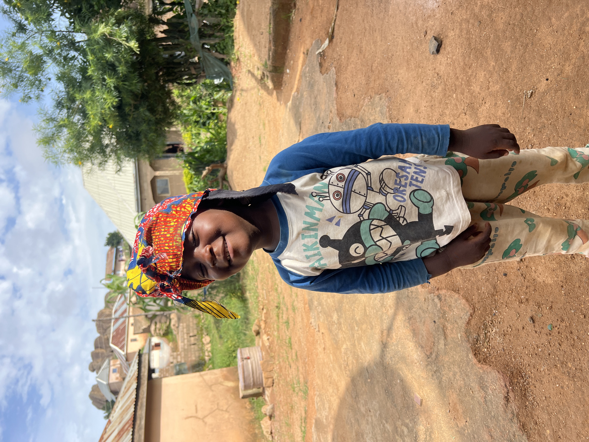A young child with dark skin and closed eyes is smiling at the camera in an outdoor, rural setting. The child is wearing a long-sleeved blue and white shirt with cartoon characters and colorful patterned pants. A vibrant, patterned headwrap adorns the child's head. In the background, there are several buildings with corrugated metal roofs, some scattered rocks, and lush green vegetation, including trees and what appears to be a corn stalk. The ground is predominantly reddish-brown dirt and some paved areas. The overall impression is a candid portrait of a child in a simple, everyday environment. The location is the city of Jos, Nigeria.
