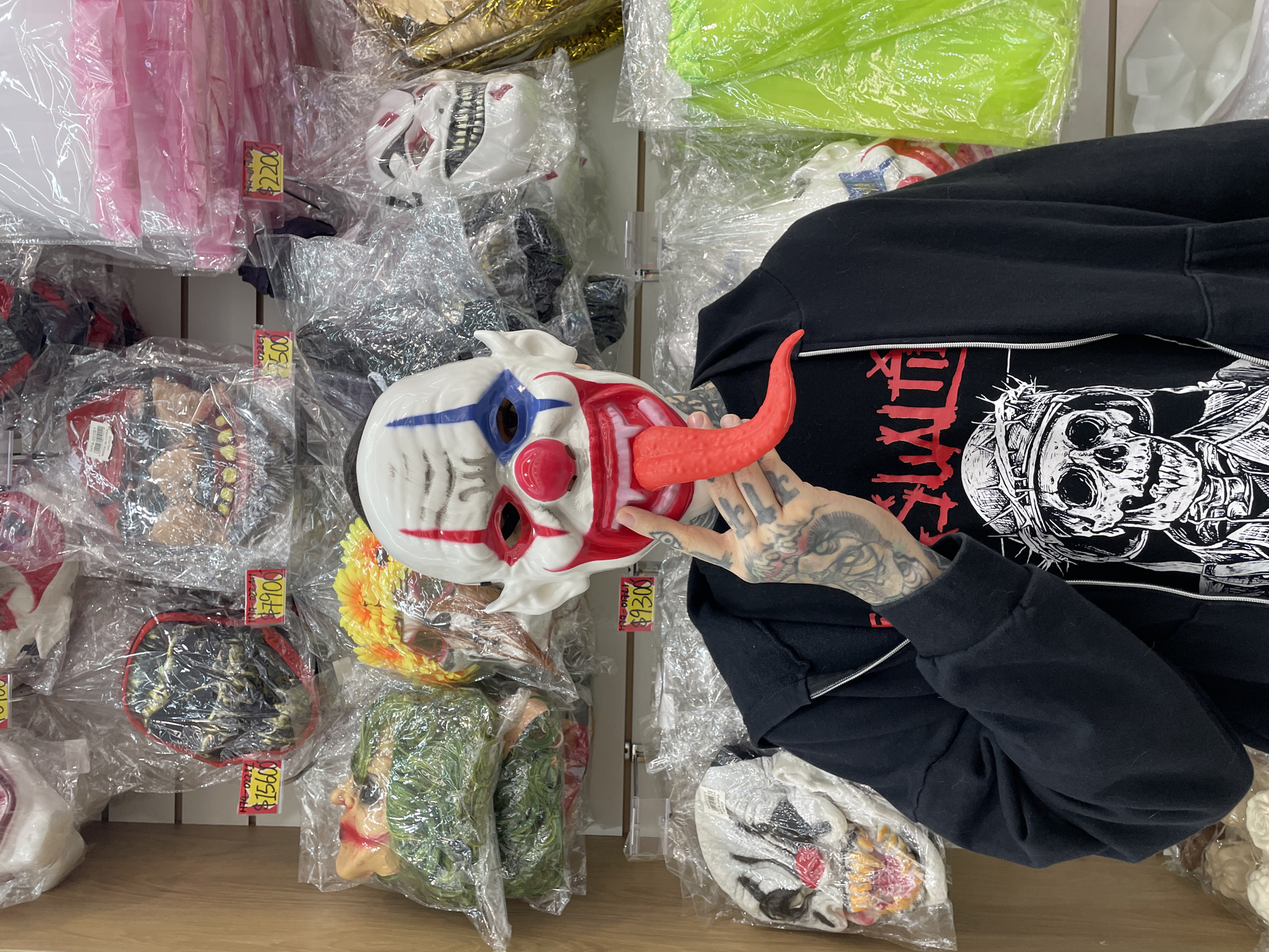 In what appears to be a shop or market stall in Buenos Aires, Argentina, a person is captured holding a distinct white clown mask with blue stripes, red cheeks, and a large, red, protruding tongue. The individual wears a black zip-up hoodie over a black t-shirt. The t-shirt features a bold graphic, partially visible, including a detailed black and white illustration of a skull adorned with a crown of thorns, and some red text that appears to include "XIII" and other characters. The person's left forearm is extensively covered with black ink tattoos, depicting various intricate designs, including what resembles a goat or ram's head.
The background reveals a display of numerous other masks and costume accessories, each typically sealed in a clear plastic bag and hung or stacked on white shelving units. These items include a white and red monster-like mask, a green-haired mask, and a yellow, floral-themed mask. Other items, possibly costumes or accessories, are packaged in bright pink and neon green plastic. Several red and yellow price tags are visible, indicating prices in what are likely Argentinian Pesos (ARS), given the context. Visible prices include "$2200," "$2500," "$1900," "$1500," and "$9300." The scene suggests an indoor retail environment focused on party supplies, Halloween costumes, or novelty items.