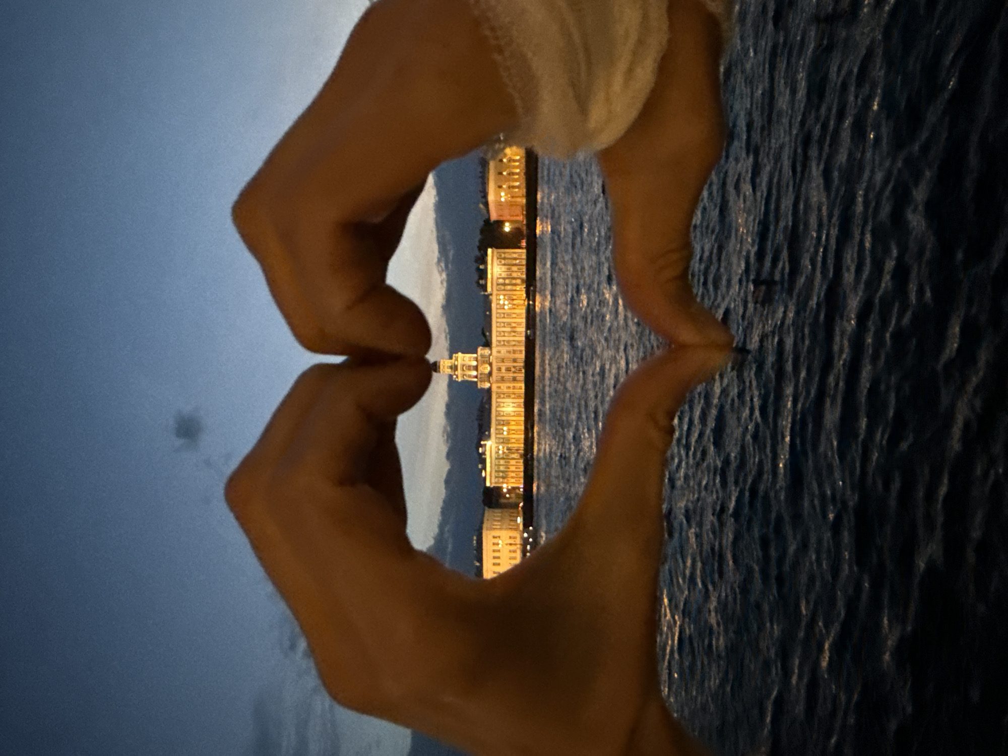 The image, oriented in landscape, presents an evening or night scene in Saint Petersburg, Russia. Two human hands in the foreground create a heart shape, framing the cityscape beyond. The hand on the right side features a light-colored bandage or fabric wrapped around the wrist.
Through the heart, a line of historically styled buildings is prominently illuminated with warm yellow lights, contrasting against the dark twilight sky. A distinctive central building with a prominent architectural feature, possibly a dome or spire, stands out among the structures across the water. The body of water, likely a river or canal, occupies the mid-right portion of the frame, appearing dark with subtle ripples and reflections of the city lights.
The overall impression conveys a sense of affection or appreciation for the iconic architecture of Saint Petersburg at night, as expressed by the heart gesture. There is no visible text in the image.