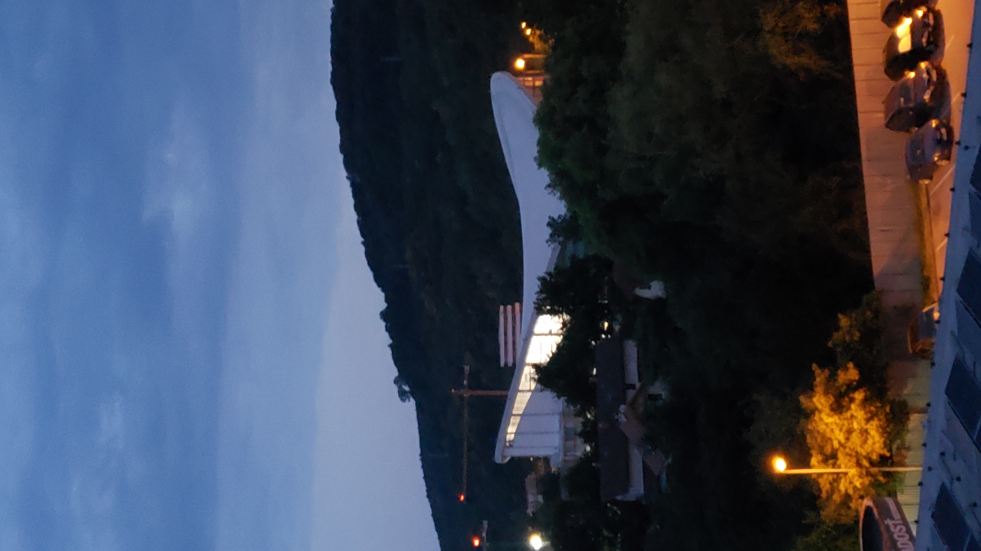 This image, taken at dusk in Prešov, Slovakia, depicts a view of a modern building with a distinctive white, curved roof, possibly a stadium or arena, situated on a tree-covered hillside. The building is illuminated from within, casting a warm glow. In the foreground, a street lamp illuminates a tree with bright yellow leaves, and to the left, a portion of a structure with the word "DOST" visible is present. Further in the foreground, a parking lot is visible with several cars parked. The sky is a deep blue with scattered clouds, and a construction crane is visible on the left side of the frame, suggesting ongoing development. The overall atmosphere is serene, capturing the transition from day to night in a semi-urban environment.