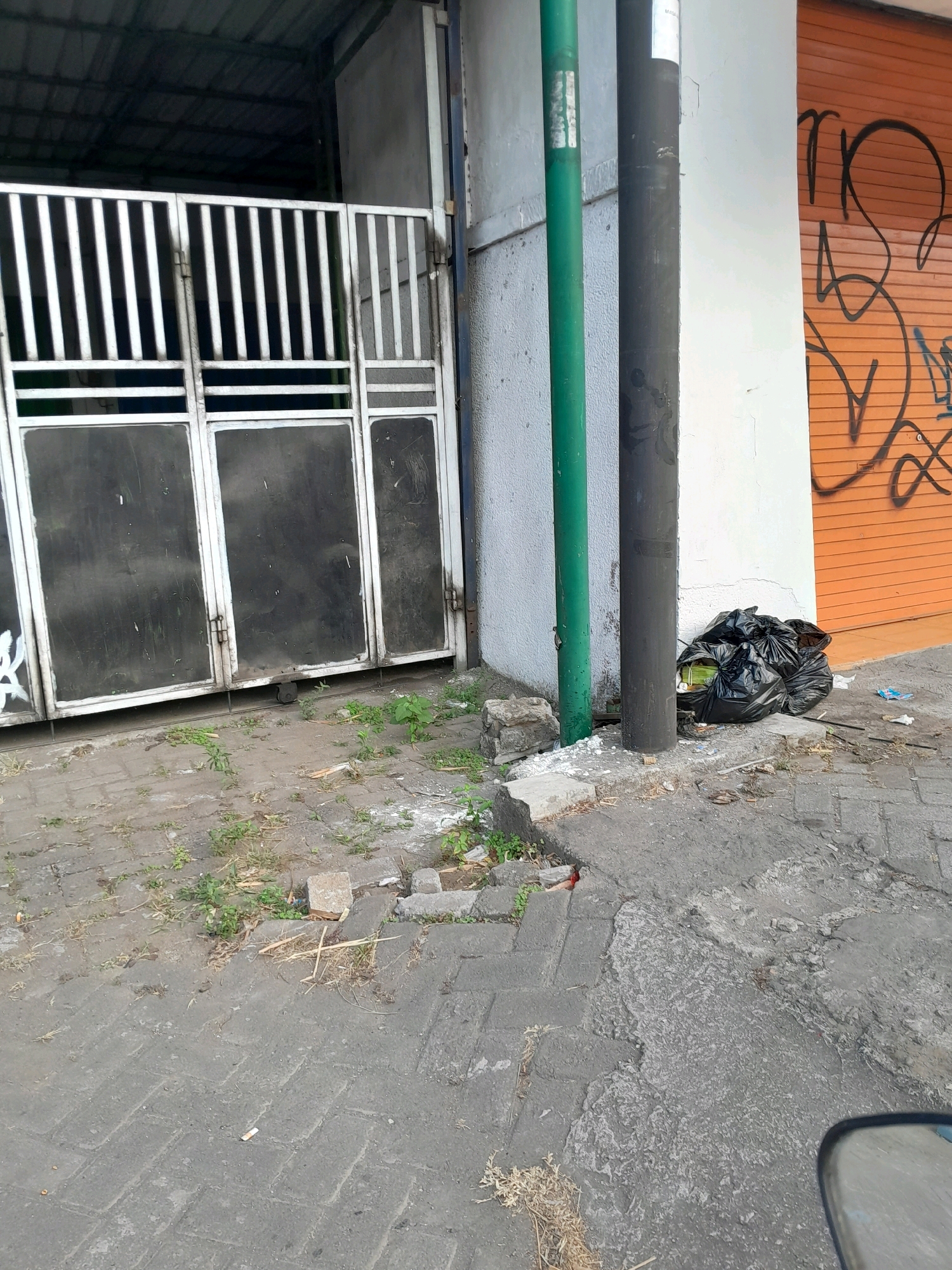 The image depicts an outdoor scene in Pakisaji, Indonesia, focusing on a section of a building's exterior and the adjacent walkway. In the background, a white building wall is visible, with an orange rolling door covered in black graffiti on the right side. Two pipes, one green and one black, stand vertically near the wall. To the left of the pipes, a white metal gate with vertical bars and horizontal supports is partially open, revealing a dark interior space with a corrugated metal roof. In front of the gate, a paved walkway made of interlocking concrete blocks shows signs of wear and tear, with several patches of dirt and weeds growing between the blocks. A pile of black garbage bags sits on the walkway near the base of the pipes, with some visible contents that appear to be organic waste. Scattered debris, including small stones and dried plant matter, is present on the ground. The overall scene suggests an urban or semi-urban environment, possibly a commercial or industrial area, with elements of neglect and untidiness.