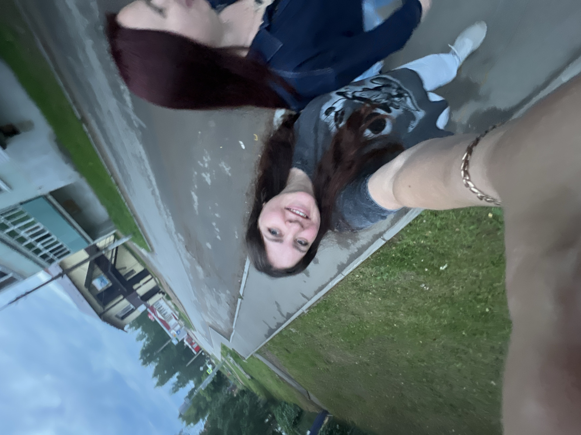 This is a selfie taken by two young women outdoors in Noviy, Russia. They are standing on a wet paved surface, likely after rain, with a grassy area and some buildings visible in the background. The image is rotated 90 degrees counterclockwise.
The woman in the foreground is looking directly at the camera with a smile. She has long, dark brown hair and is wearing a gray t-shirt with a graphic print. A gold chain bracelet is visible on her right wrist.
The second woman is partially visible in the background. She has long, dark red hair and is wearing a dark denim jacket over a blue garment. Both women appear to be in casual attire, suggesting a relaxed outing.
The scene suggests a cloudy day, given the dull lighting and the wet pavement. The presence of buildings and a road indicates an urban or suburban environment. The overall mood is cheerful and candid, characteristic of a casual selfie. No visible text is present in the image.