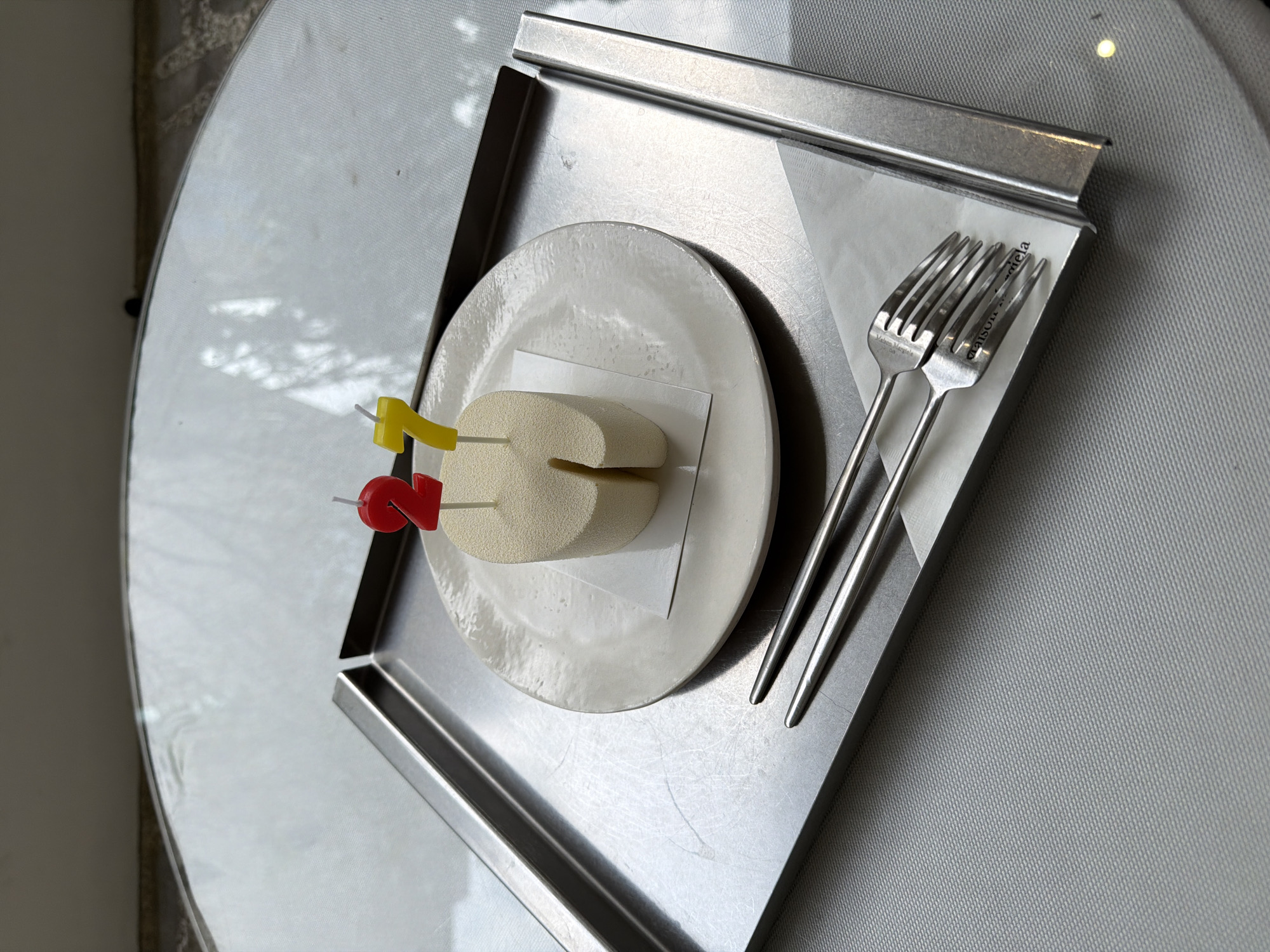 The image displays a minimalist dessert presentation, likely for a birthday celebration, set indoors in Seoul, South Korea. A small, light-beige cylindrical dessert, possibly a cake or mousse, featuring a vertical cut, is centered on a textured white ceramic plate. Embedded in the dessert are two birthday candles: a red number "2" and a yellow number "7," signifying a 27th birthday.
The plate rests on a piece of white parchment paper, which is placed within a rectangular, brushed metal tray. Alongside the plate, two polished silver forks lie on the parchment paper. The brand "MAISON Margiela" is distinctly engraved on the handles of both forks.
This metal tray is situated on a reflective, round table, possibly made of glass or a similar material, which reflects the surrounding light and shadows. The scene is illuminated by bright, natural daylight, indicated by the shadows and light patterns on the table and the softly blurred background. The setting appears to be a contemporary and clean indoor environment, with no people visible. The overall mood suggested is celebratory and understated.