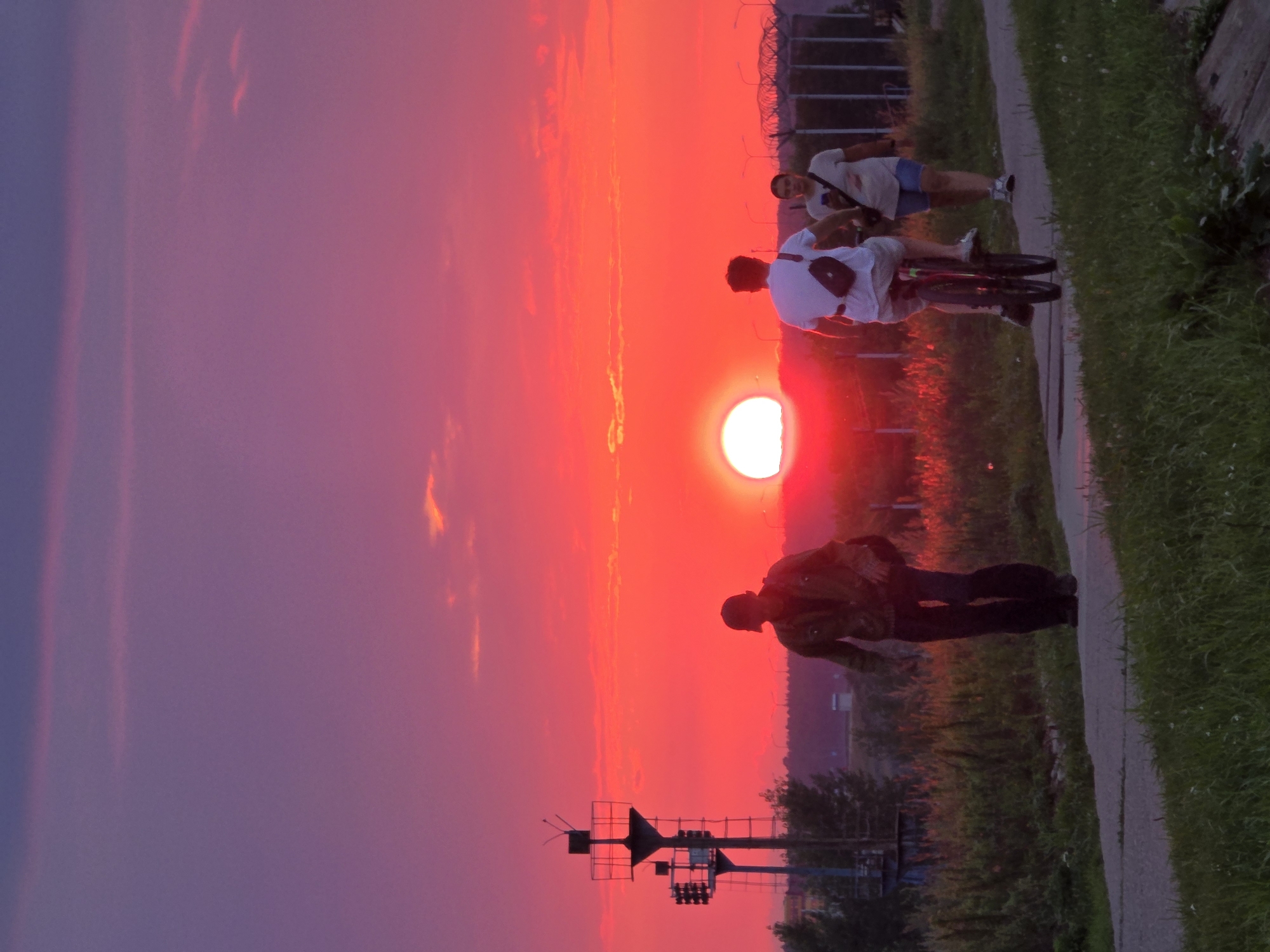 A silhouette of a man walks along a paved path as the sun sets with a vibrant red and purple sky. He wears a dark jacket, dark pants, and a cap. Behind him, a person rides a bicycle, and another person walks. The cyclist is wearing a white t-shirt and shorts, and the walker is in a light-colored t-shirt and blue shorts. In the background, a tall industrial structure with lights stands to the left, and a fence with lights runs along the right side of the path. The path is bordered by green grass and vegetation. The scene appears to be a waterfront path in Chaikovsky, Russia, likely near the Kama River dam.