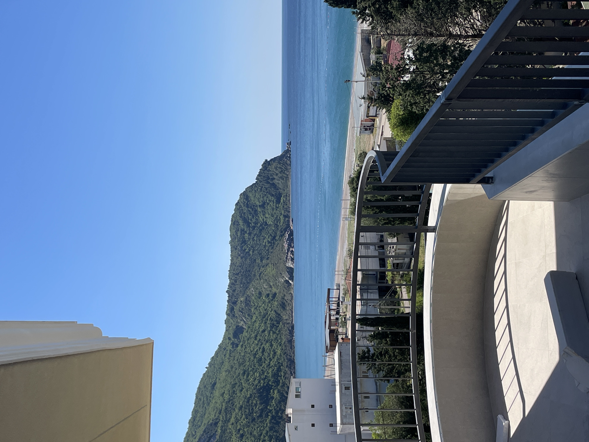 The image presents a serene coastal landscape in Čanj, Montenegro, captured from an elevated viewpoint, likely a balcony. In the foreground, a dark grey metal railing of a modern balcony curves across the bottom right, with distinct, long shadows cast onto the light-colored tiled floor by the bright sunlight. Parts of the building's light-toned facade are visible on the left and right edges.
The midground showcases the vast, calm blue sea, extending horizontally across the frame. Along the coastline on the right, a narrow sandy or pebbly beach is visible, with several low-rise buildings and scattered green trees behind it. Small white buoys or markers are spaced out in the sea, possibly indicating a swimming area. To the left, a large, densely forested green mountain or hill slopes down to meet the water, forming a rugged and lush natural boundary.
The background consists of a clear, bright blue sky, devoid of clouds. The overall scene suggests a sunny day with excellent visibility, likely mid-morning or early afternoon given the angle and intensity of the shadows. There are no people or discernible text visible in the image. The setting conveys a peaceful and scenic vacation atmosphere.