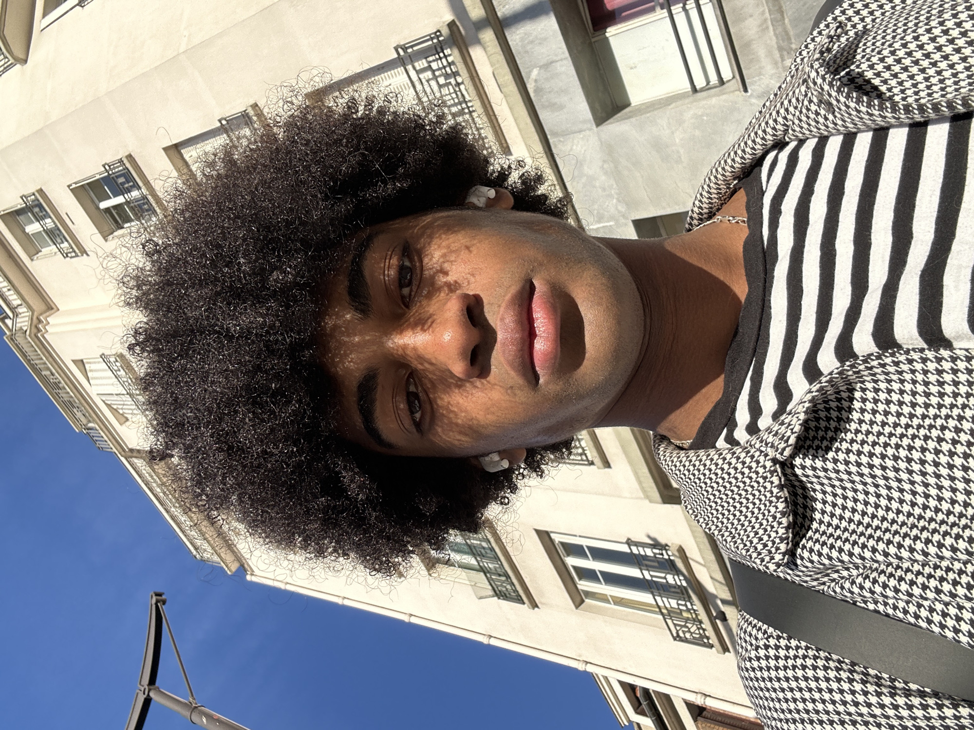A low-angle shot features a person with an Afro hairstyle, fair skin, and full lips. The person is wearing what appears to be a striped shirt under a black and white houndstooth patterned jacket. A black strap is visible over the houndstooth. Sunlight casts shadows on the person's face, obscuring details. The person has white earbuds in their ears.
In the background, a light-colored building with multiple windows stretches upwards. The sky is a clear blue. The architecture suggests an urban environment, potentially Paris, France. A black metal pole is visible near the bottom left corner of the frame.