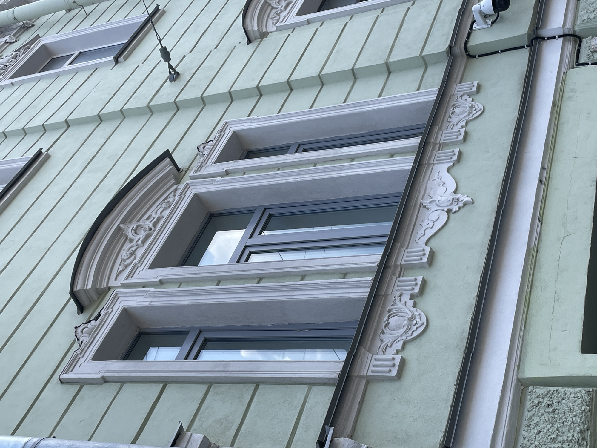 The image shows a portion of a building facade in Saint Petersburg, Russia. The building has a light green painted exterior with horizontal, slightly angled panels. Three windows are visible, each with ornate white frames and multiple panes. The middle window has a curved, decorative frame at the top. A white security camera is mounted near the top right of the image. A white drainpipe runs vertically down the facade, partially obscuring one of the window frames, which is decorated with ornate white plasterwork.