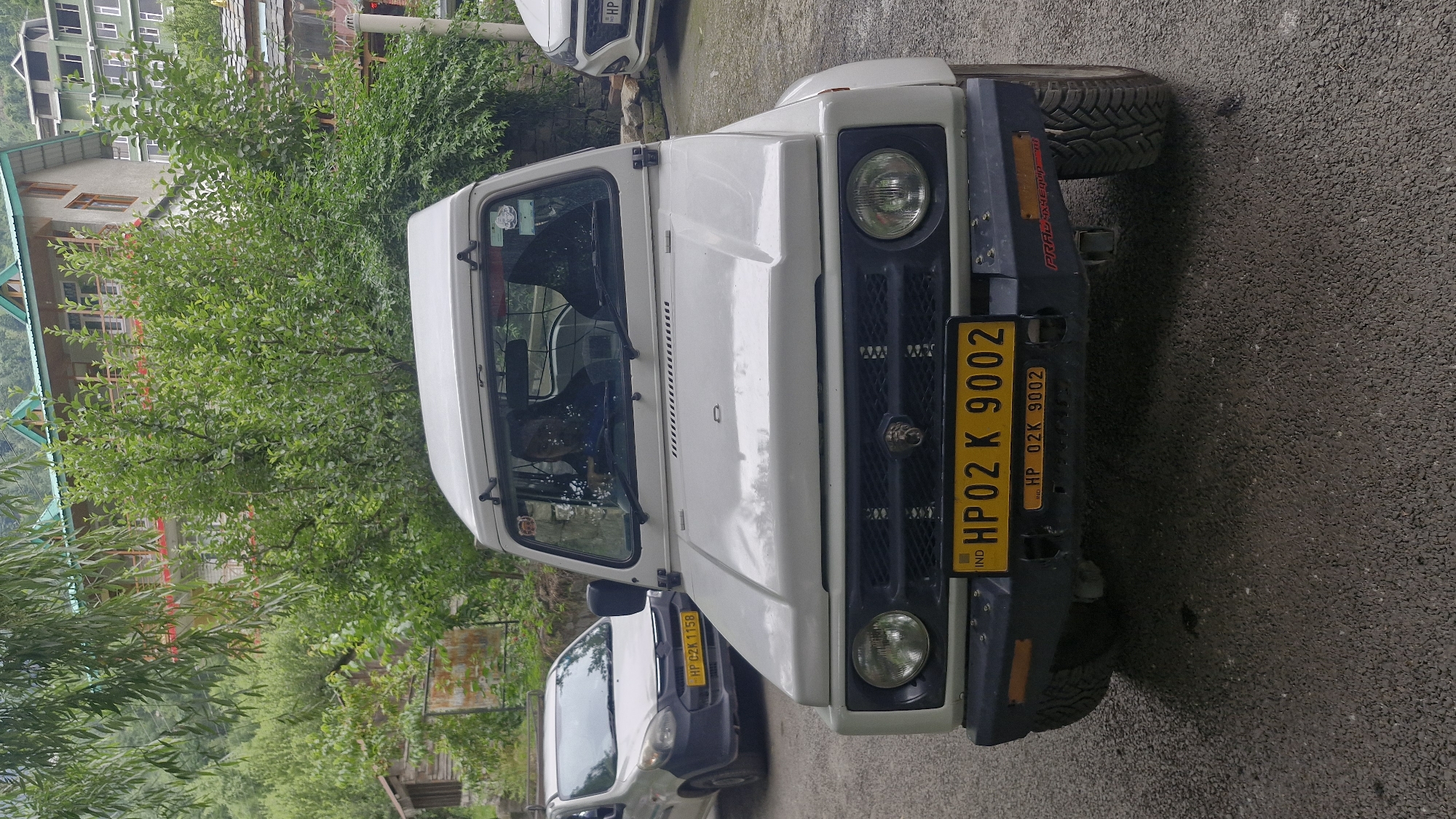 A white Suzuki Gypsy is parked on a paved road in Vashisht, India. The vehicle's front is facing the camera. A yellow license plate on the front of the vehicle reads "HP02 K 9002". A smaller yellow sticker below it says "HP 02K 9002". The vehicle has a black grille with a silver emblem in the center and two round headlights. The windshield is visible, and a person is seated in the driver's seat, wearing a blue shirt. To the left of the Gypsy, a silver car is parked. In the background, trees and buildings are visible. One building, likely a hotel, has a traditional Himachali architectural style with a sloping roof. The sky appears overcast.