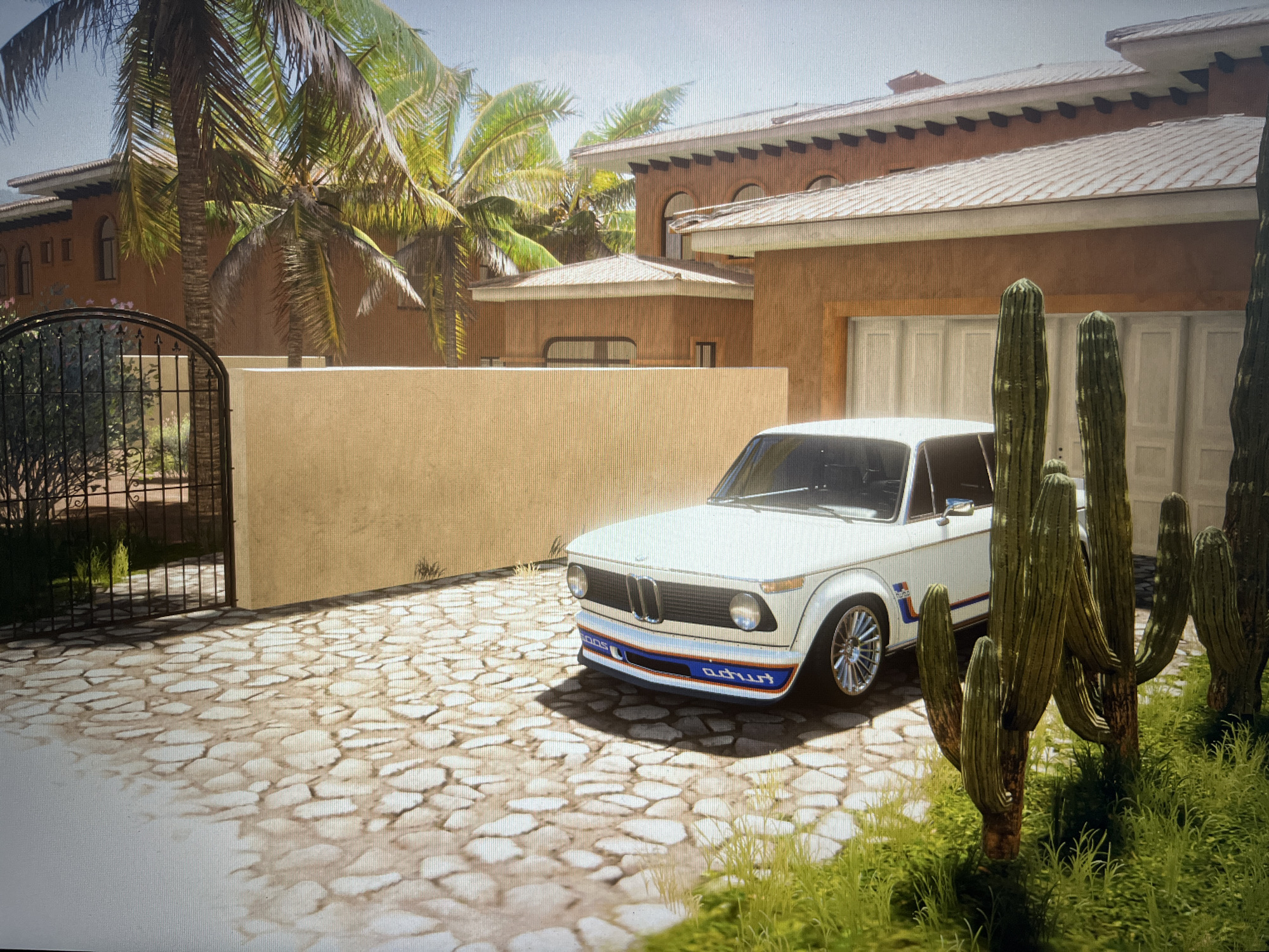 The image depicts a white vintage BMW 2002 Turbo parked on a cobblestone driveway in front of a large, modern villa. The villa has terracotta-colored walls and a tiled roof, with arched windows and a wide garage door. Palm trees and cacti are visible in the surrounding landscape, suggesting a warm climate. The car is facing the viewer, with its front bumper displaying racing stripes in blue, white, and red, and the word "turbo" visible. The driveway is illuminated by sunlight, casting shadows from the car, cacti, and a black wrought-iron gate to the left of the frame. The scene appears to be a still from a video game, likely Forza Horizon 5, given the graphical style and the presence of a virtual car in a stylized environment. There are no people present in the image. The location of Varna, Bulgaria is not depicted or implied by any elements within the image.