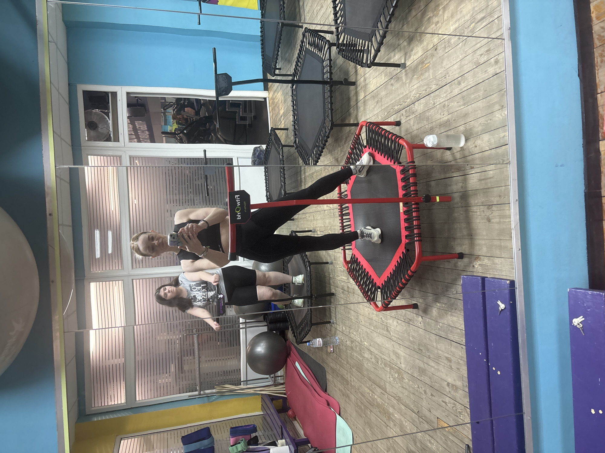 A medium shot shows the reflection of a young woman, likely Caucasian, who is wearing black athletic wear, in a gym setting. She is standing on a mini trampoline. The reflection reveals other gym equipment, including several trampolines and exercise balls. The gym is indoors with light blue walls and wooden floors. A water bottle rests on one of the trampolines. The exact location cannot be determined from the image.