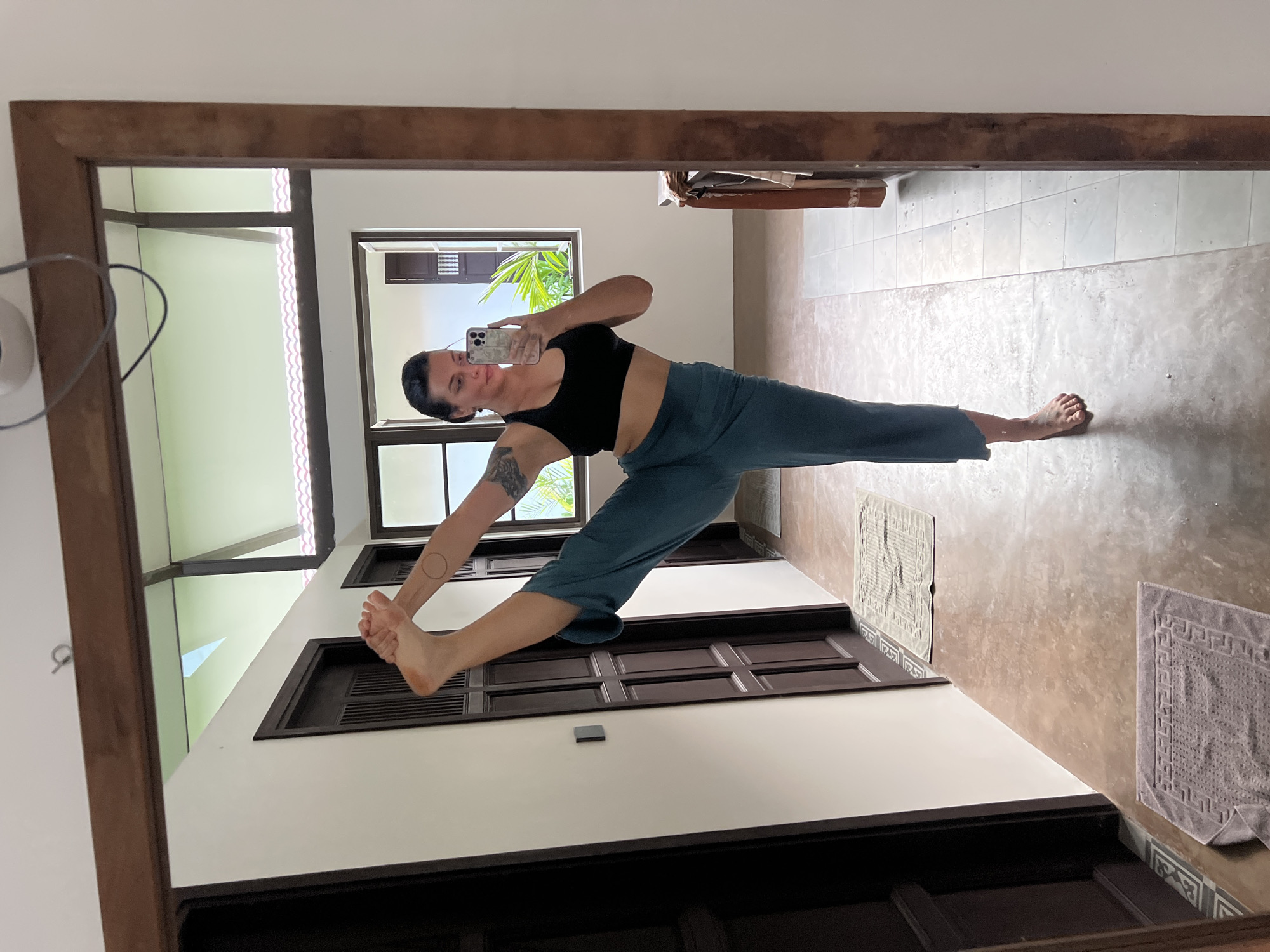 A woman is taking a selfie in a mirror. She is in a yoga pose, with one leg extended forward and the other bent behind her. She is wearing a black sports bra and teal leggings. She has dark hair pulled back and a tattoo on her left arm. The background appears to be an indoor space with large windows and wooden doors. The flooring is tiled. The overall setting suggests a home or a studio, possibly in a tropical location given the palm fronds visible through the window. The image seems to be taken during the daytime, with natural light coming in from the windows. There are no visible signs of text in the image.