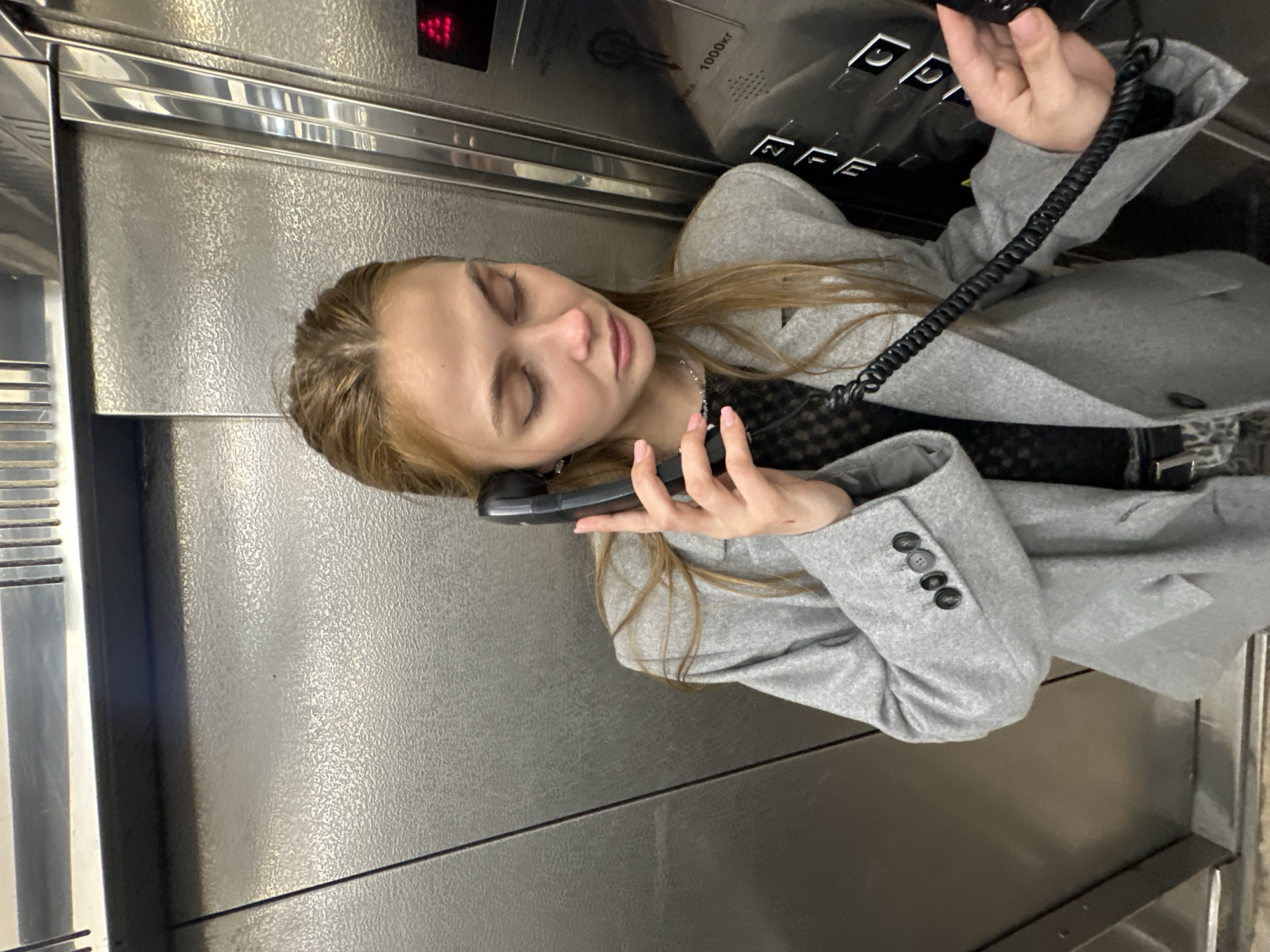 A young woman with light brown hair is in an elevator, holding a black telephone receiver to her ear. She is wearing a grey coat over a black top. The elevator has a stainless steel interior and a black telephone with a coiled cord attached to the receiver. The elevator's floor indicator shows a red "8". The city of Krasnoyarsk, Russia, is not visually discernible in this image.