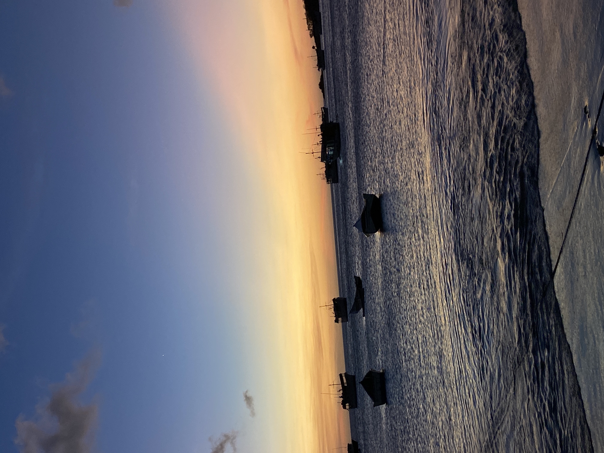 The image depicts a coastal scene in Juan Griego, Venezuela, during twilight, characterized by either sunrise or sunset. The sky transitions from deep blue at the zenith, with a single bright point of light in the upper left, through lighter blue to a prominent band of yellow and orange above the horizon, incorporating subtle pink coloration in the upper right. Scattered dark gray clouds are present in the upper left quadrant. The sea surface occupies the lower two-thirds of the frame, exhibiting small ripples and waves, reflecting the ambient light. Approximately a dozen vessels of varying sizes are distributed across the water. These include at least five larger, dark-silhouetted boats with masts and superstructures, consistent with fishing or utility vessels, primarily located in the mid-ground. Several smaller, open skiffs are positioned closer to the foreground and mid-ground. Dark, undulating land masses are visible as silhouettes on the far right horizon. The immediate foreground shows a dark, wet, flat surface, consistent with a concrete pier or embankment, at the water's edge. No people or active interactions are visible.