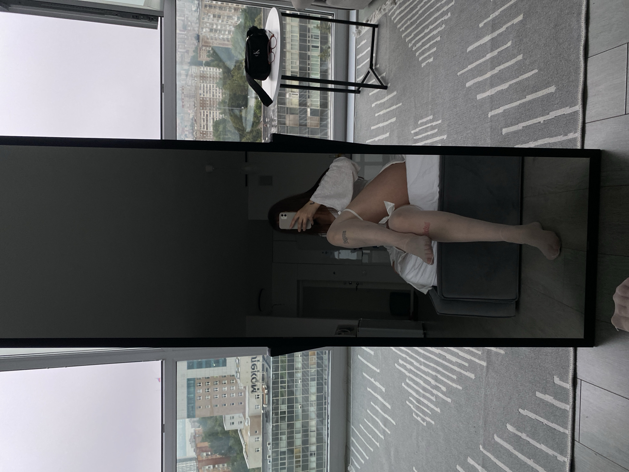 A full-length mirror reflects a young woman sitting on a bed in what appears to be a modern apartment. The woman, dressed in a white top and sheer white stockings, is holding a smartphone and taking a selfie. She has tattoos on her legs. The room features a large window with a view of a city skyline, indicative of an urban setting. Buildings of various architectural styles and heights are visible, along with a grey, overcast sky. A small round table with a black fanny pack and glasses sits near the window. The floor is covered with a grey rug with a striped pattern. The overall impression is of a contemporary living space with an urban backdrop.