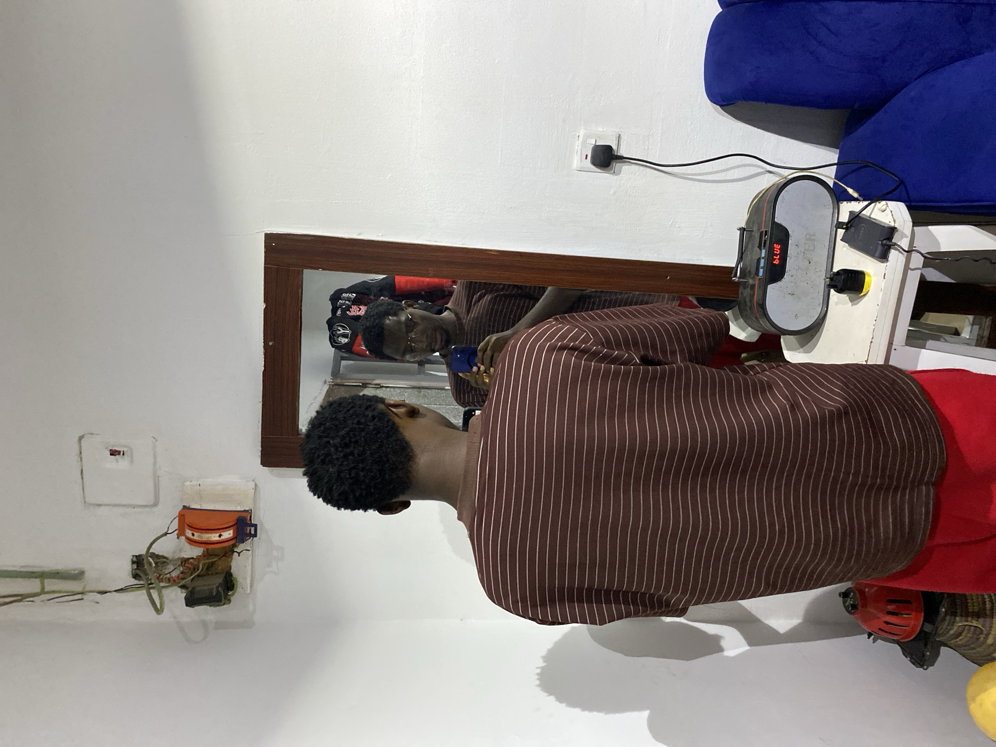 The image shows a young man inside a room, captured from a side profile view, facing a mirror. He has short, dark, curly hair and wears a brown long-sleeved shirt with thin white vertical stripes, and red pants. His reflection in the rectangular mirror, framed in dark wood, shows him looking directly forward, wearing dark-rimmed glasses, and holding a blue smartphone. He appears to be taking a photo or video of himself.
The room features plain white walls. To the right of the mirror, a white wall socket is visible with a black plug and cable connected, which leads to a grey and black electronic device. This device, possibly a portable speaker or power bank, sits on a small white stand and displays "BLUE" and "30.19" in red digital text. Another cable from this device leads towards a partial view of a blue upholstered sofa or chair arm in the upper right background.
To the left of the mirror, an older wall-mounted electrical installation with exposed wires, an orange component, and a white square cover above it is present. On the floor to the far right, a red and black object, resembling part of a machine or fan, and a yellow object are visible. The setting is an unadorned indoor space, likely a living area or bedroom. The image lacks specific time-of-day indicators. The overall impression is a personal moment of self-documentation within a functional, but not elaborately decorated, room in Jos, Nigeria.