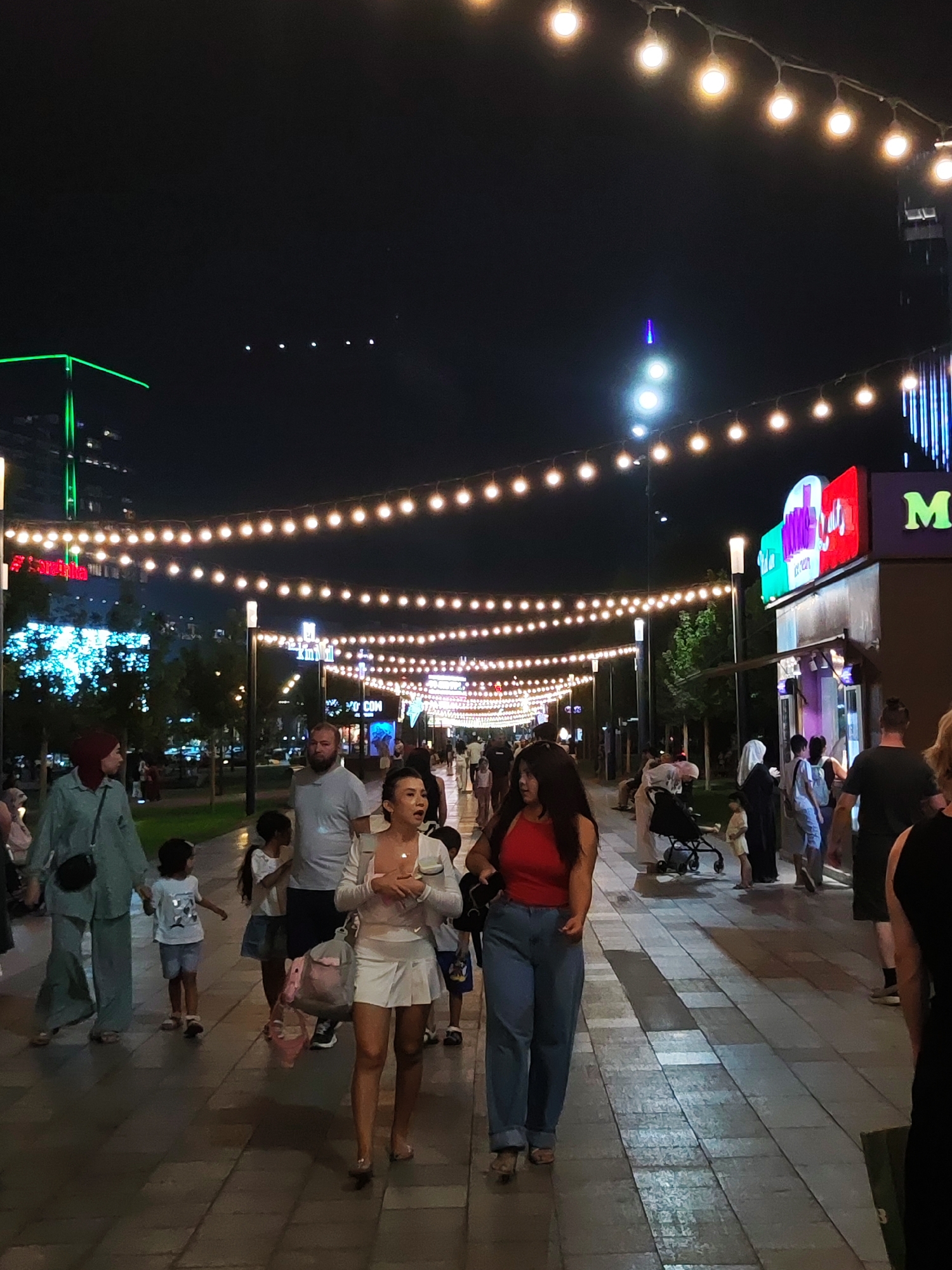 The image depicts a pedestrian area at night, illuminated by strings of lights, in Tashkent, Uzbekistan. The scene appears to be a lively street or promenade with various people walking and interacting. In the foreground, a woman in a white top and skirt is walking, holding a bag. To her left, a woman wearing a hijab and a mint-colored outfit is walking with a young child. Further down the pathway, other individuals, including families with children and couples, are visible. Businesses with illuminated signs, including one with "M" and colorful graphics, are present on the right side of the image. The background features buildings, trees, and more strings of lights extending into the distance. The overall atmosphere is that of a well-lit, active urban space at night.
