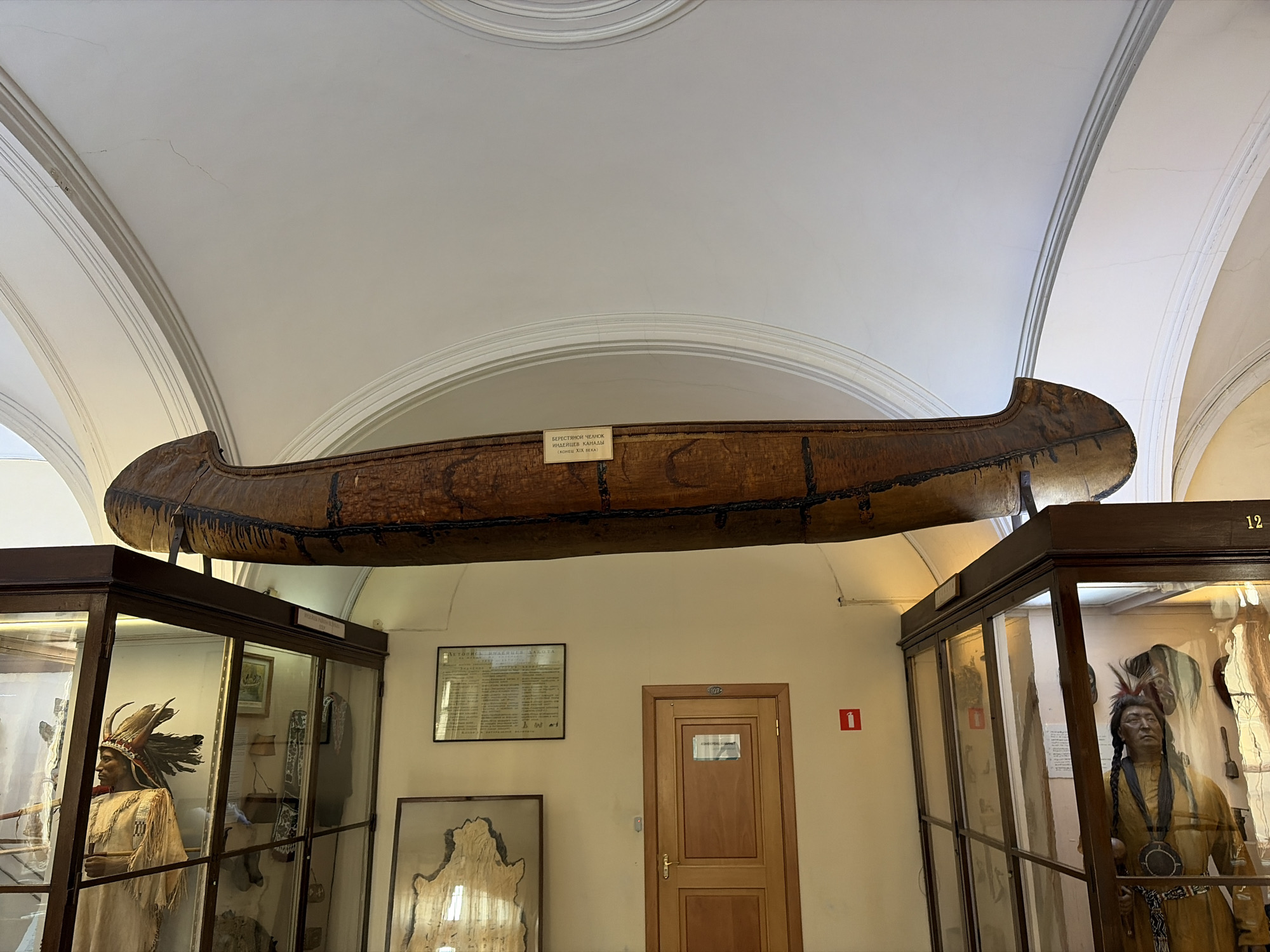 The image shows an interior view of a museum, likely in Saint Petersburg, Russia, based on the Cyrillic writing on a plaque attached to the canoe. A large, dark brown canoe is suspended from the ceiling, above two glass display cases. The display cases contain artifacts, including mannequins dressed in Native American clothing and headdresses. A closed wooden door is centered in the background with two framed documents and one red fire extinguisher sign on the wall, behind and to the left of the door. The room features a high, arched ceiling with decorative moldings. The lighting appears to be a mix of natural and artificial light. The overall impression is that of a historical or ethnographic museum exhibition, possibly focused on Native American culture.
