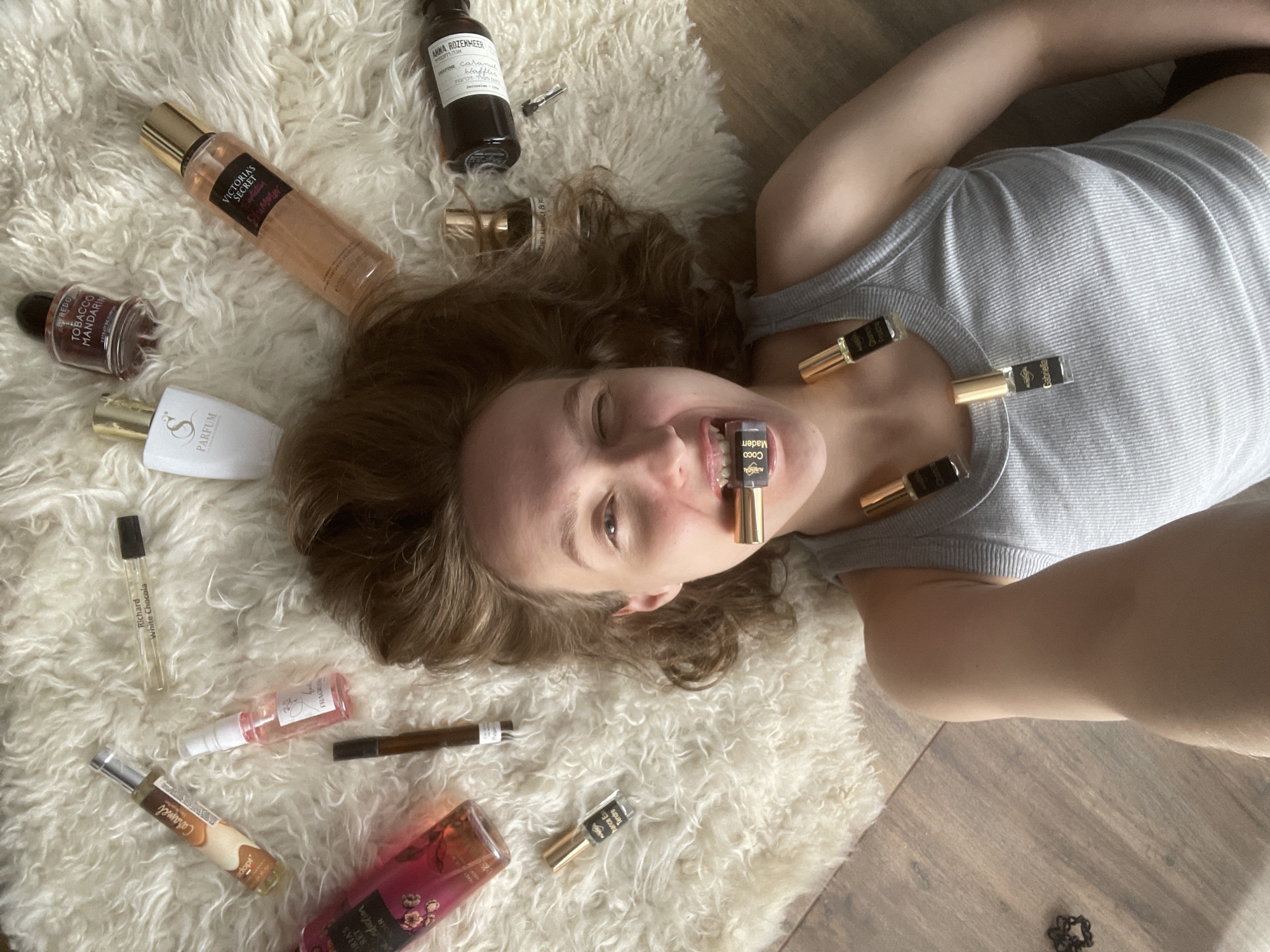 A woman lies on a fluffy white rug, surrounded by numerous perfume bottles of various sizes and shapes. Several small perfume vials are positioned on her chest and held in her mouth. The rug is partially on a wooden floor. The brand names "Victoria's Secret", "Anna Rozekmeer", "Byredo", "Richard White Chocola", and "S Parfum" can be read on some of the bottles. The location "Подшивалово, Russia" is not indicated in this image.