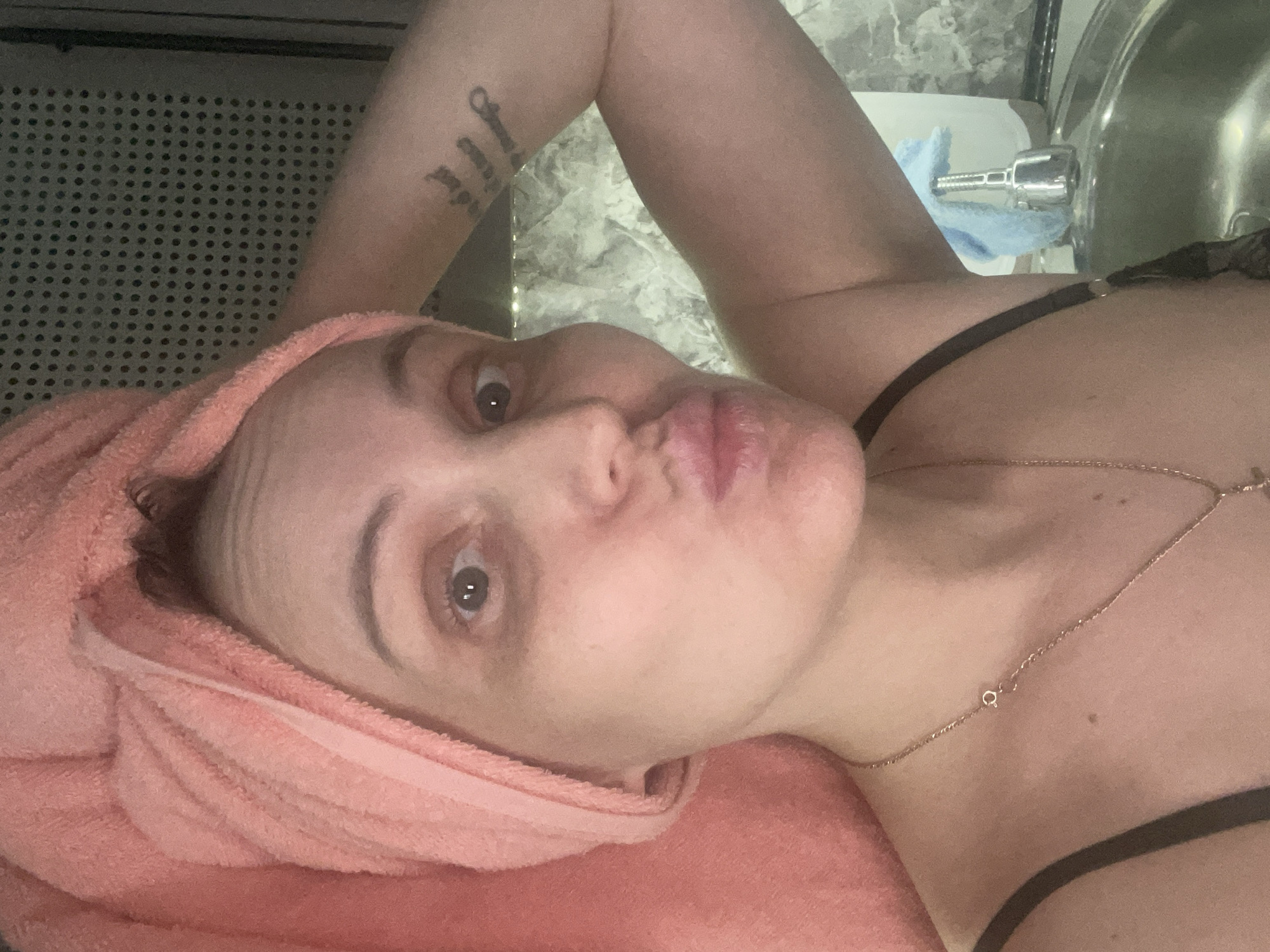 A woman with a pink towel wrapped around her head is lying down. She has a tattoo on her arm. She is wearing a black bra and a gold necklace. Behind her is a sink and a faucet with a blue towel. To her left is a textured surface with small holes.
There is no evidence in this image to suggest the city of Chaikovsky, Russia is relevant.