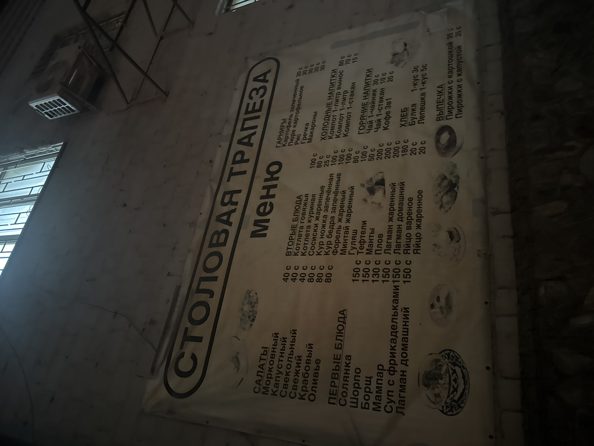 The image shows a large, beige banner with a black border, titled "СТOЛOВАЯ ТРАПEЗА" (Canteen Feast). The banner displays a menu with various food and drink items and their prices in Russian. The menu is divided into sections such as "САЛАТЫ" (Salads), "ПEРВЫЕ БЛЮДА" (First Courses), "ГАРНИРЫ" (Garnishes), "ХОЛОДНЫЕ НАПИТКИ" (Cold Drinks), "ГОРЯЧИЕ НАПИТКИ" (Hot Drinks), "ХЛЕБ" (Bread), and "ВЫПEЧКА" (Baked Goods). Some of the items listed include "Морковный" (Carrot) salad, "Солянка" (Solyanka) soup, "Картофель запеченный" (Baked potato), "Компот" (Compote), "Чай" (Tea), "Кофе" (Coffee), "Булка" (Bun), and "Пирожки с картошкой" (Pirozhki with potato). There are also small, stylized illustrations of food items on the banner. The banner is attached to a rough, grey, concrete wall. Above the banner, a metallic grate covers a window. To the right of the window, a wall-mounted air conditioning unit is visible. The overall scene suggests an outdoor food establishment or a public canteen, likely in Bishkek City, Kyrgyzstan, given the context of the Cyrillic alphabet and common dishes. The lighting is dim, indicating it might be evening or a poorly lit indoor space. There are no people visible in the image. The banner is slightly creased and appears to be weather-worn.