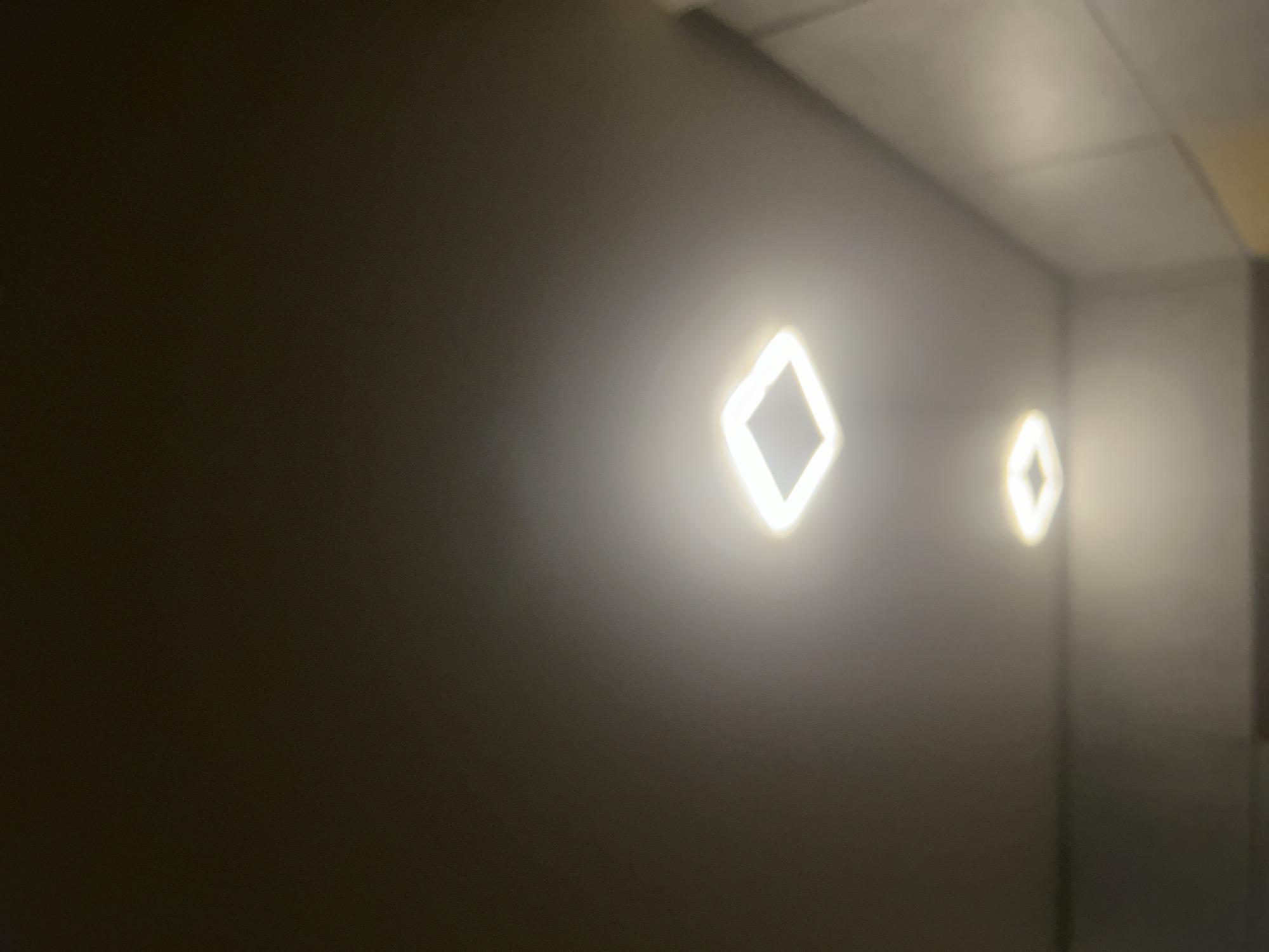 The image shows a dimly lit interior space with two diamond-shaped light fixtures mounted on a dark wall. The wall appears to extend into a corner. The ceiling above is visible, suggesting an enclosed room or hallway. The area is sparsely furnished.
