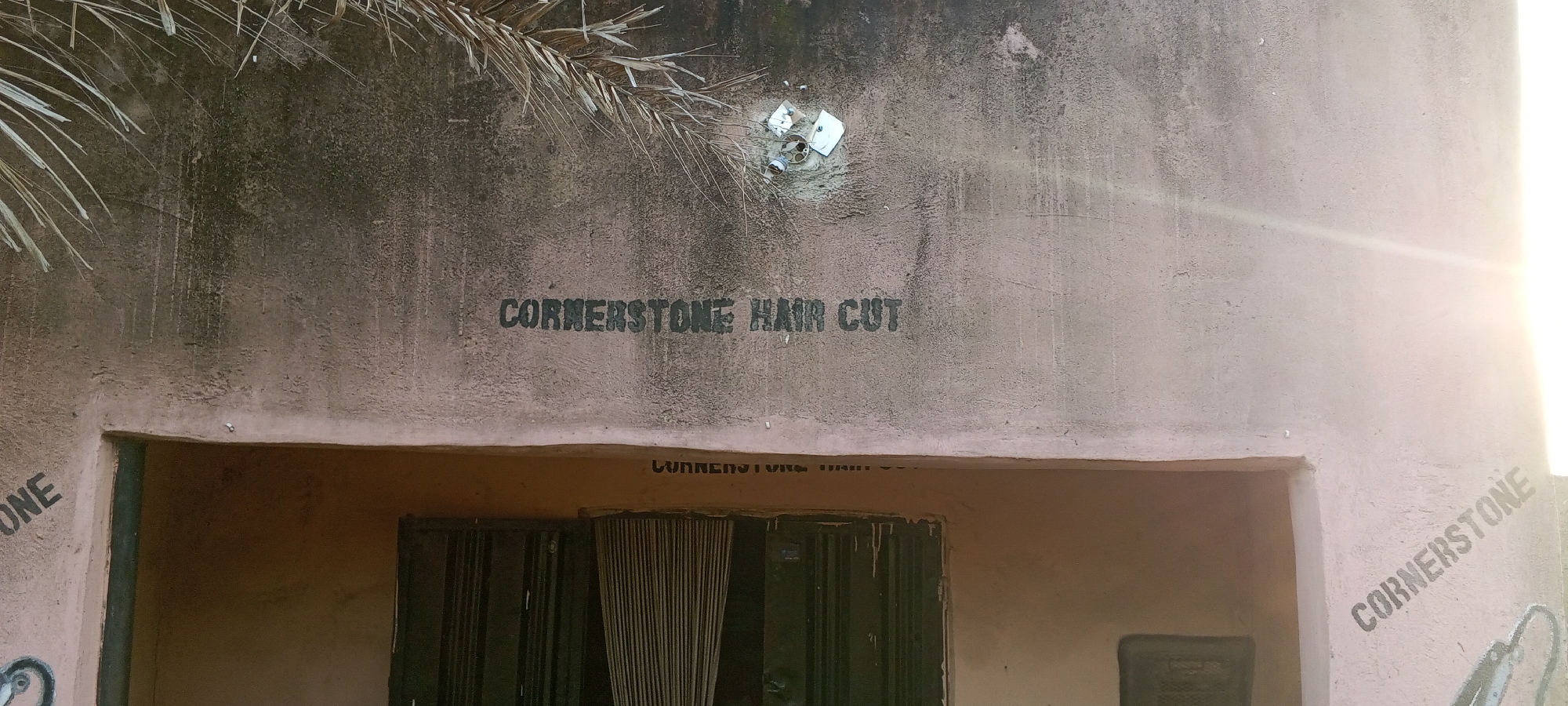 The image shows a faded sign on a weathered, pinkish-brown building, likely in Jalingo, Nigeria. The sign reads "CORNERSTONE HAIR CUT" in stenciled, dark lettering. Above the sign, a dry palm frond hangs from the upper edge of the building. To the right of the sign, sunlight flares intensely on the wall. Below the main sign, another faint, possibly older, stenciled word is partially visible. The entrance to the building features dark, closed doors, with vertical blinds visible through a gap in the center. To the left of the entrance, the word "ONE" is visible, also stenciled. Further to the right, the word "CORNERSTONE" is stenciled diagonally on the building's exterior. The overall impression is of an aged establishment, possibly a barber shop or salon, situated in a warm, possibly dry climate.