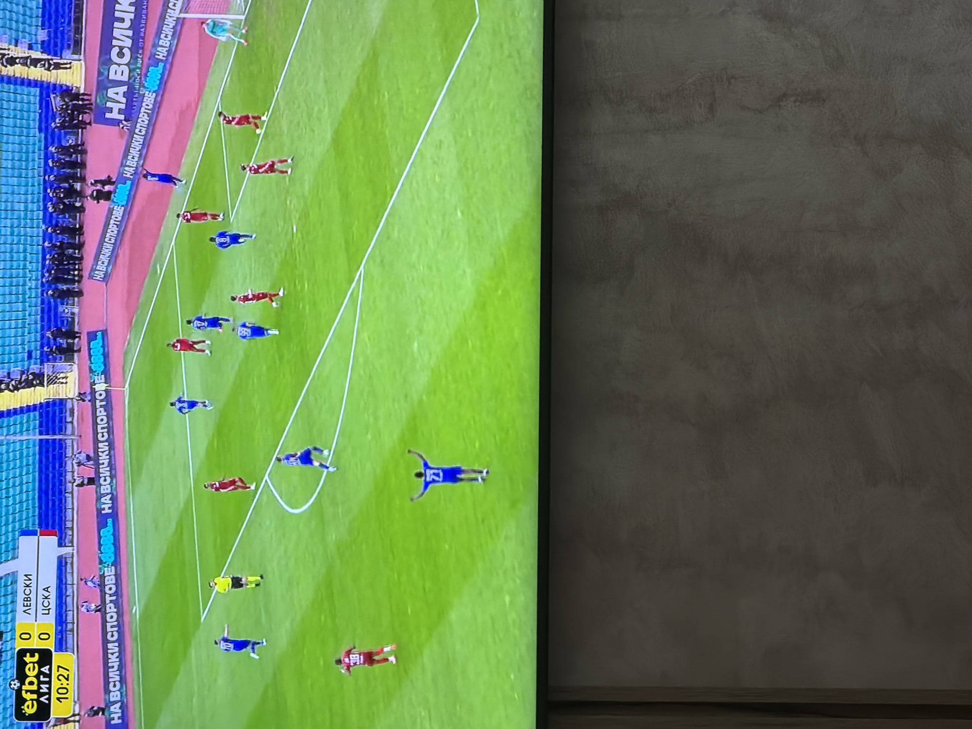 A close-up, slightly blurred, high-angle shot shows a television displaying a soccer game. The game appears to be a Bulgarian league match between Levski and CSKA, with the score at 0-0 and the time at 10:27. The game is taking place at a stadium where the advertisements, "НА ВСИЧКИ СПОРТОВЕ" (for all sports) are displayed in blue. The city location, Plovdiv, Bulgaria cannot be determined solely from the image.
The focus is on the field of play, where players in blue and red uniforms are spread across the green grass. A referee in yellow and black stripes is also visible. The angle suggests a high-angle view of the game, possibly a wide shot taken from a camera positioned in the stadium.
The background shows a gray textured wall.