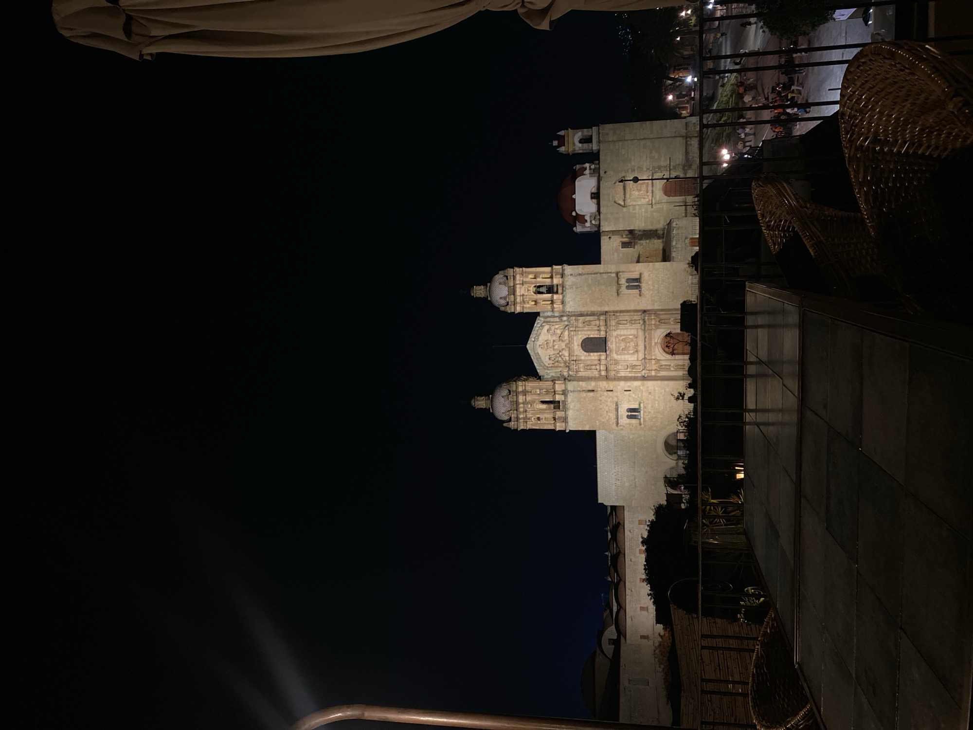 The photograph, taken at night, depicts a building with architectural details illuminated against a dark sky. It appears to be an outdoor scene, possibly from a balcony or elevated location, suggested by the presence of table and chairs. In the background, there are multiple people gathered. This scene is located in the city of Oaxaca City, Mexico.