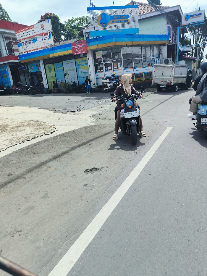 The scene is a street in Ciwidey, Indonesia. A woman wearing a hijab is riding a motorcycle. The motorcycle is black with a blue license plate. There is a white dividing line on the street. The street is paved and has a few potholes. On the left side of the image, there are several buildings. One building has a sign that says "Apotek al mazoem". There are other people walking and riding bicycles. There is a truck parked on the right side of the image. The sky is clear and blue. The street is mostly empty.