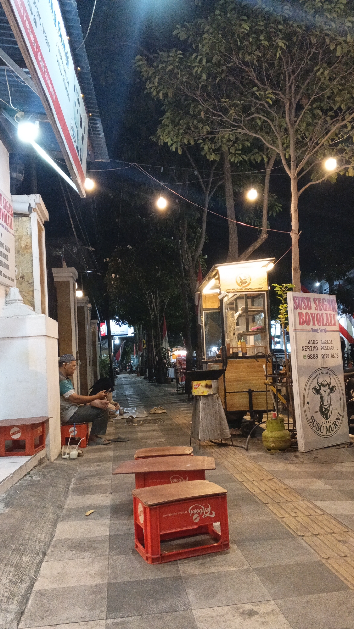 The image depicts a street scene at night in Bojonegoro, Indonesia. On the left, a man is sitting on red plastic crates, looking at his phone. In front of him are two red plastic crates used as tables. Further down the street, a mobile food cart is visible, illuminated by overhead lights. A sign on the cart reads "SUSU SEGAR BOYOLALI." Several trees line the sidewalk, and string lights are hung between them. The sidewalk is paved with checkered tiles. The overall impression is that of a lively, outdoor food stall area at night.