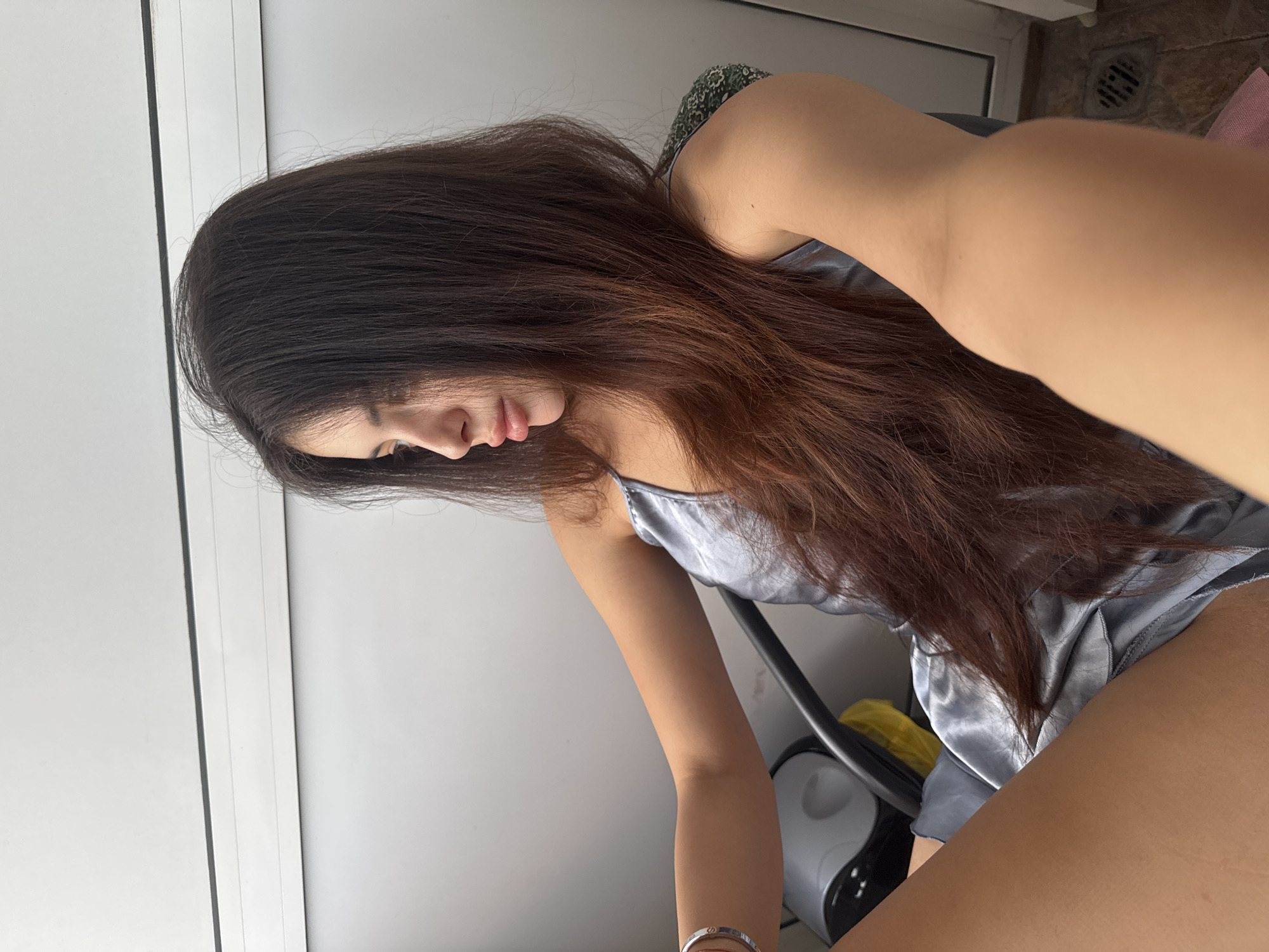 The image captures a young woman from a slightly elevated perspective, seated and looking downwards and to her left. Her long, dark brown hair, with noticeable reddish-brown highlights towards the ends, flows over her shoulders and down her back. She is wearing a light grey or silver satin camisole top. A silver bracelet adorns her left wrist. Her lips are slightly parted, and her expression appears calm and contemplative.
The background on the right side of the frame features vertical white panels, possibly part of a door or wardrobe. To the left, behind her, a textured wall, appearing to be made of stone or rough material, is visible, along with a dark circular vent. A dark chair provides support. Below her left arm, a grey and black appliance, possibly a fan or air purifier, is partially seen, with a yellow item behind it. The lighting is bright and natural, indicating it is daytime. No discernible text is visible within the image. The location context for this scene is Plovdiv, Bulgaria.