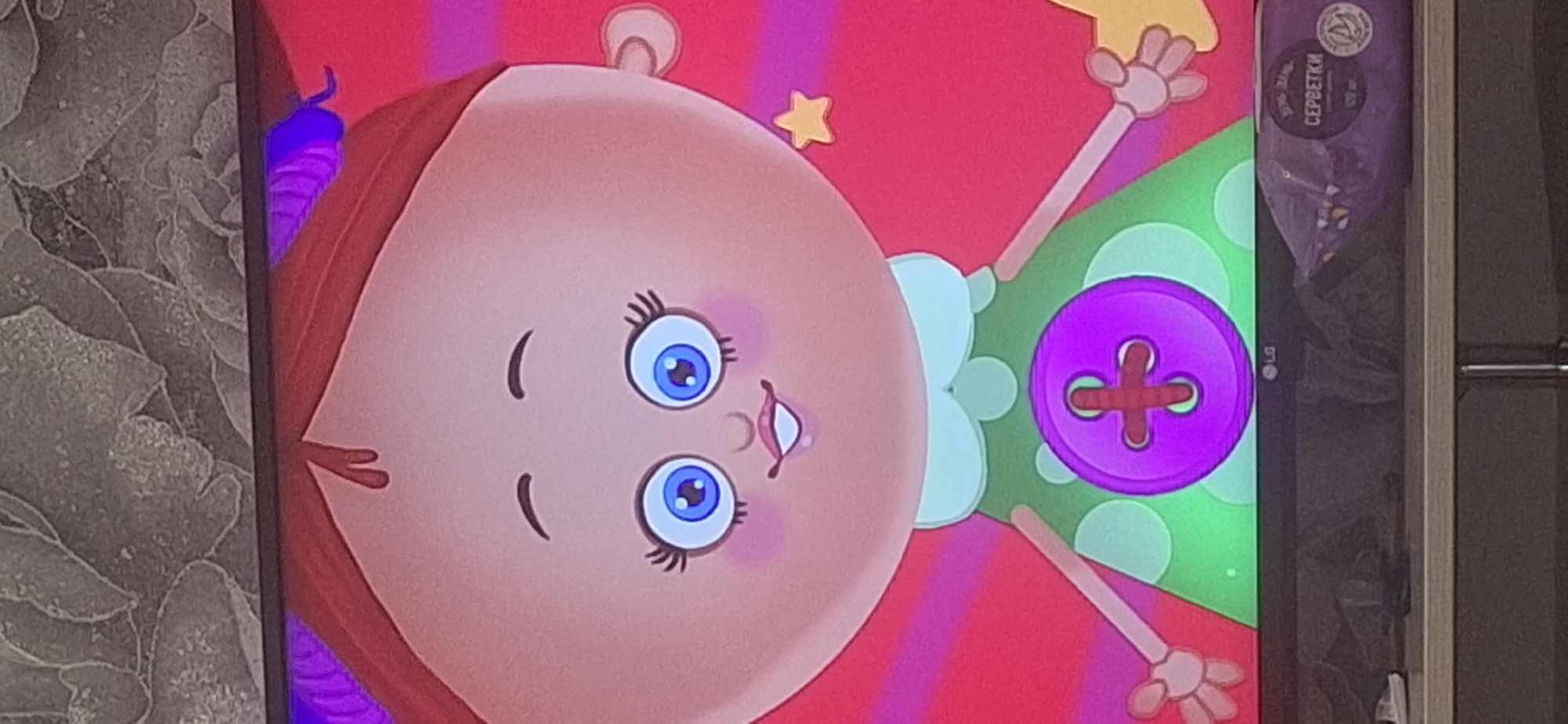 The image shows a close-up of a cartoon character, a doll-like figure with a large, round head, blue eyes, and a wide smile. The character is wearing a green polka-dot dress with a large purple button in the center. The background is a bright pink and yellow gradient with radiating lines. The doll is depicted in what appears to be a drawing or animation, with a simplified style. The bottom of the image shows the top of a black LG television screen, with a shelf below it holding some items, including a package of wipes with Russian text. The overall impression is of a bright, cheerful, and whimsical scene, possibly from a children's program. There is no indication of any specific location in Bila Tserkva, Ukraine.