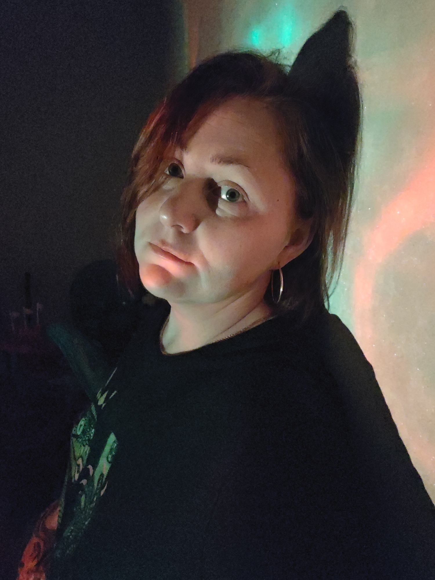 A woman is positioned in a dimly lit indoor space. She has reddish-brown hair, fair skin, and is wearing a black shirt and silver hoop earrings. The background wall features a diffused light pattern with green, white, and red hues. A dark, indistinct object, possibly a cat ear headband, is visible atop her head. The location of the scene in Markovskii, Russia, is not evident from the image.