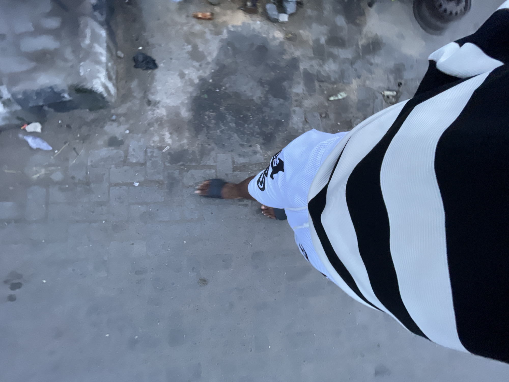 A first-person perspective shot shows a person walking on a paved surface in Lekki, Nigeria. The person is wearing a black and white striped long-sleeved shirt and white shorts with a black logo. The ground is dirty with some puddles and scattered debris, including plastic bags and bottles.