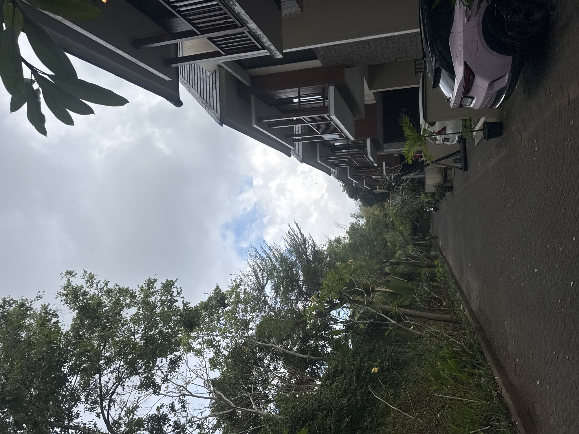 The image depicts a pathway lined with lush greenery on the left and a row of modern, multi-story buildings on the right. The pathway appears to be paved with dark interlocking bricks and is empty except for a motorcycle visible in the distance on the left side. The buildings on the right have balconies with wooden railings and large windows. A light pink car is parked in front of one of the buildings on the right. The sky is overcast with patches of blue visible. The overall impression is a well-maintained, tropical residential or resort area in Kuta Selatan, Indonesia.