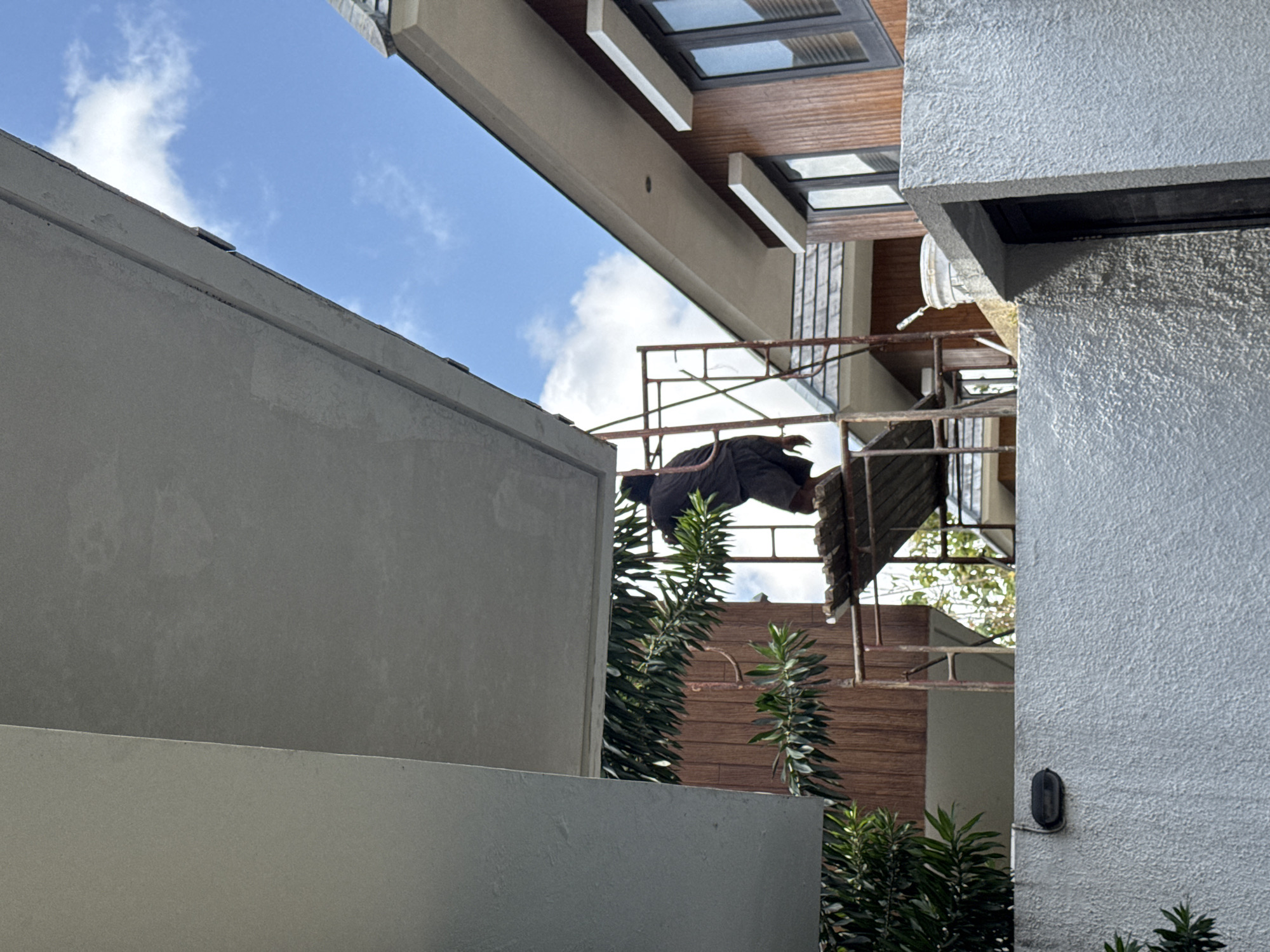 In Kuta Selatan, Indonesia, a construction worker is seen on scaffolding, applying material to the overhang of a building. The worker, dressed in dark clothing and shorts, is positioned near the edge of the overhang. The scaffolding, made of metal poles, supports wooden planks that form a platform for the worker. Behind the worker, the building features brown wood paneling and dark-framed windows. The sky above is blue with scattered white clouds. In the foreground, a textured gray wall is visible with a black outdoor light fixture attached. Lush green foliage from a tree partially obscures the view of the worker and the scaffolding. Another building with a peaked roof is visible in the mid-ground, also appearing to be under construction or renovation. A white bucket, possibly containing building materials, sits on the edge of the wall near the scaffolding.