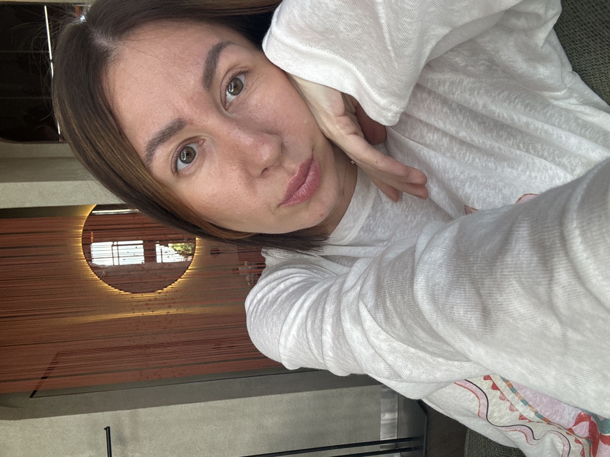 An adult female is captured in a close-up selfie in an indoor setting in Усады, Russia. Her head is slightly tilted, with her left hand resting against her jawline, fingers splayed. She has medium brown hair, hazel eyes looking directly at the camera, and a natural complexion with some subtle skin texture and slight redness on her nose and cheeks. Her lips are softly parted, revealing their natural pink color. She wears a loose-fitting, long-sleeved white top made of a textured, lightweight fabric, showing multiple folds and creases. A subtle silver-colored ring is visible on her left ring finger. A colorful, abstract pattern in red, pink, and white is partially visible on a garment or fabric in the lower left portion of the image.
The background features a decorative wall covered in numerous vertical, reddish-orange to brown strands or ropes. Centered within these strands is a circular mirror, illuminated from behind by a warm, golden glow. The mirror reflects a distinct window or balcony with vertical bars and some green foliage outside. To the far left, a dark, vertical column or frame is visible, along with a dark, horizontal bar fixture. An arched architectural opening is partially visible in the upper right background. The lower foreground shows a textured, green upholstered surface, likely a sofa or chair, on which the subject is seated or resting.