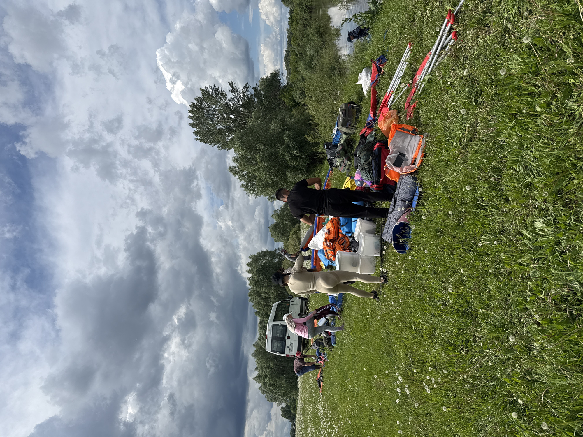A group of people are gathered on a grassy field near a body of water, possibly a river. A white van is parked nearby. They are surrounded by various camping supplies, including backpacks, buckets, and tent poles. Some individuals are standing and appear to be organizing the equipment. The sky is partly cloudy. The location is Nazyar, Russia.
