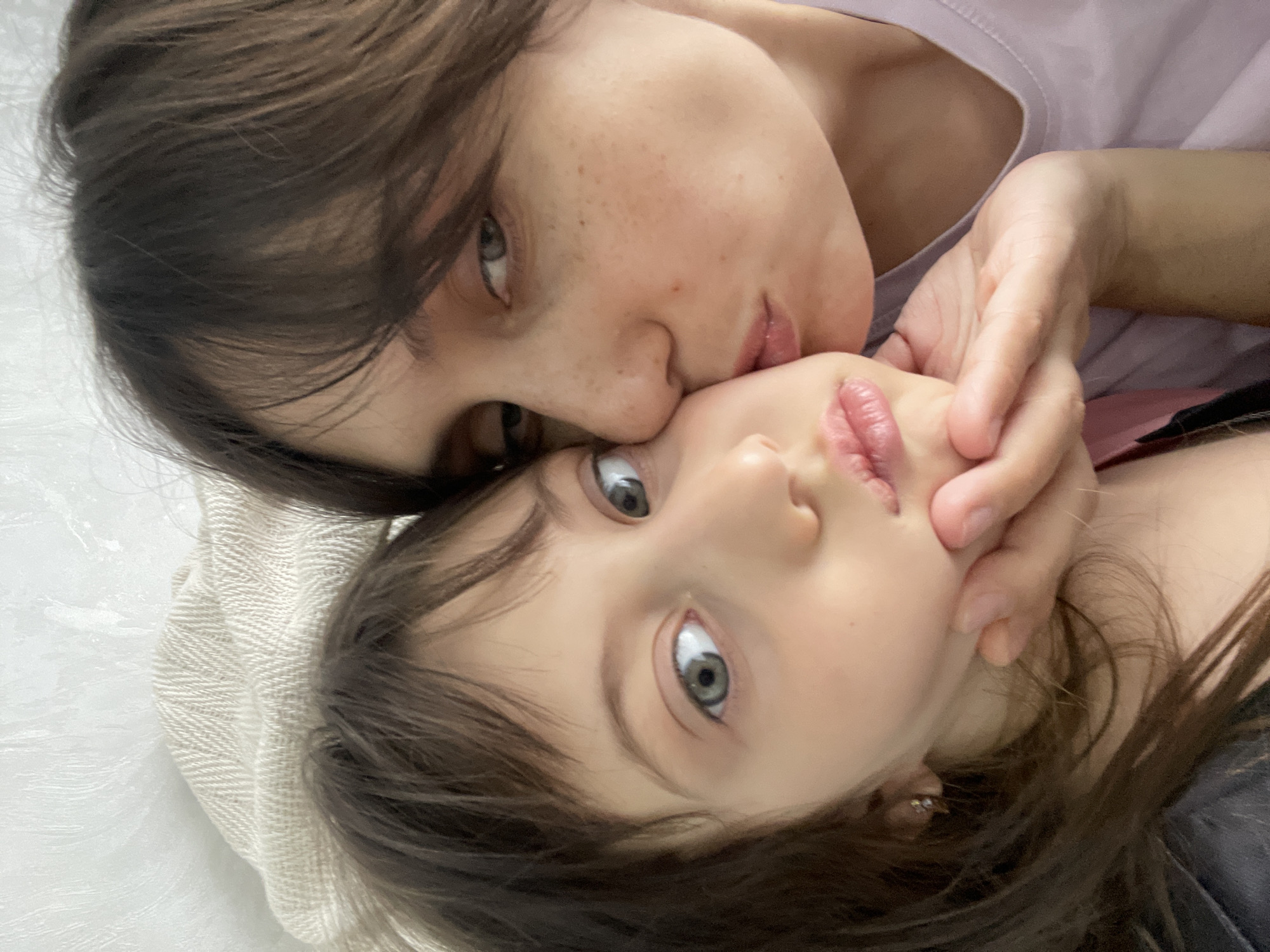 Two people are lying down close together. One appears to be an adult kissing the cheek of a child. The adult has brown hair and visible freckles. The child also has brown hair and light-colored eyes. The adult's hand is near the child's face. A light-colored, textured surface is visible in the background. There is no indication that this image was taken in Izhevsk, Russia.