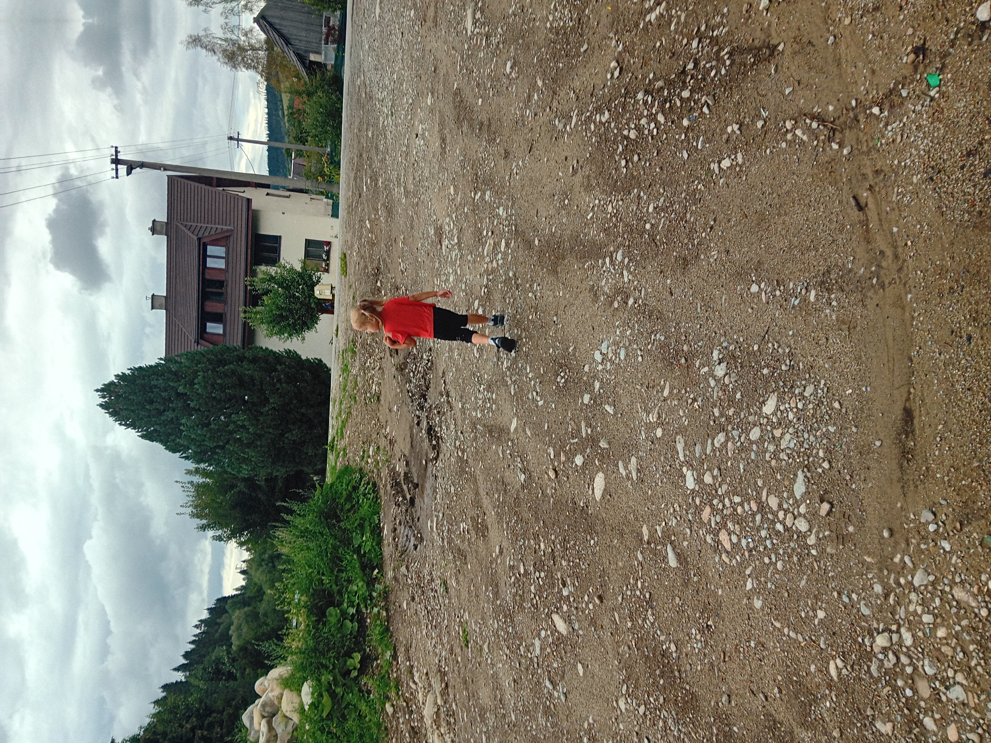 In Habovka, Slovakia, on a cloudy day, a young child with blonde hair in a red shirt and black shorts runs away from the camera across a gravel road. The road is littered with small stones and patches of dirt. To the left of the road, a large pile of rocks and dense green foliage are visible. Behind the foliage, a tall, dark green evergreen tree stands. Further back, a multi-story house with a brown roof and white siding is situated. A utility pole with wires runs diagonally across the image. To the right of the house, a smaller wooden structure is visible, and beyond that, a forested hillside under a gray sky.