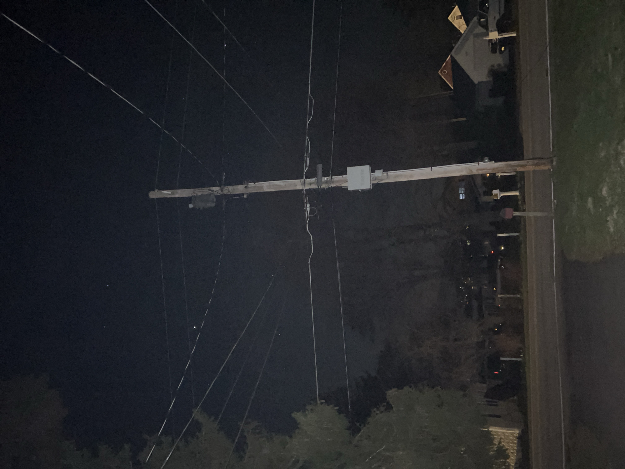 The image depicts a nighttime scene in City of Newburgh, United States of America, showing a utility pole extending across the frame from left to right. Multiple power lines are suspended in the air, creating a network of linear elements against the dark sky. The utility pole is equipped with a gray utility box and other electrical hardware. In the background, residential houses and buildings are faintly visible, interspersed with trees. A road runs parallel to the right edge of the frame, bordered by a strip of green grass or vegetation. The lighting is dim, with sparse illumination from streetlights or building lights providing minimal visibility.