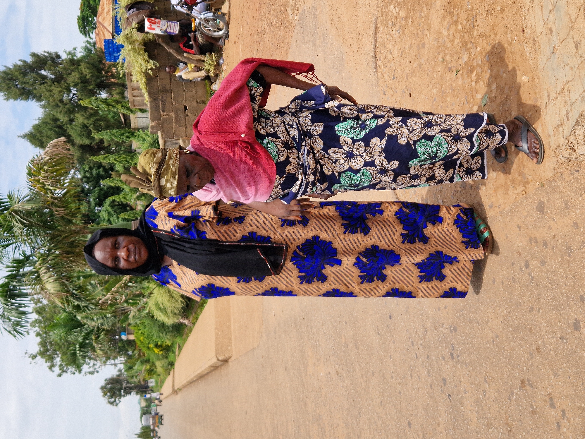 Two women stand on a dusty road in an outdoor urban environment. The woman on the left, wearing a black hijab and a long patterned dress with blue floral designs, faces forward with a neutral expression. The woman on the right, wearing a headscarf and a brightly colored floral dress with a pink shawl draped over her shoulders, looks down and to the side, appearing to lean slightly. Behind them, a street is visible with some vehicles and buildings in the distance. The ground is a mixture of dirt and cracked concrete. Vegetation, including palm trees, is present in the background.
