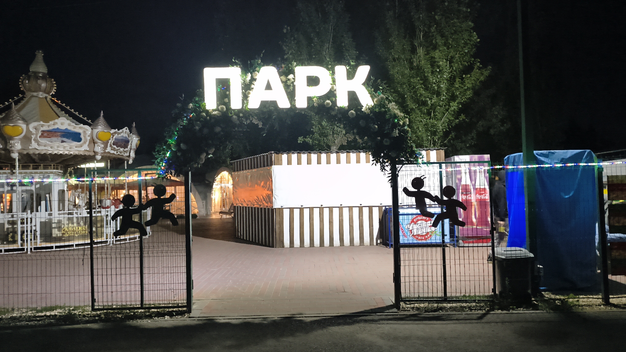 The image shows an outdoor amusement park entrance at night, in Mikhaylovka, Russia. A large, illuminated sign spelling "ПАРК" (Park) in white Cyrillic letters is prominently displayed against a dark sky, surrounded by festive lights and greenery. To the left, a carousel with ornate decorations is partially visible, with fencing and colorful lights. In the foreground, a black metal gate is open, with silhouette figures of two people on it, appearing to run. Beyond the gate, a paved walkway leads into the park.
In the center of the image, a kiosk or booth with white and brown striped walls and a large white banner is situated. To the right of this booth, there are several objects including a blue tarp-covered structure, a trash can, and vending machines with Coca-Cola branding. Trees and foliage are visible in the background, and further into the park, archways with bright lights suggest more attractions. The overall atmosphere is that of a lively entertainment venue at night.