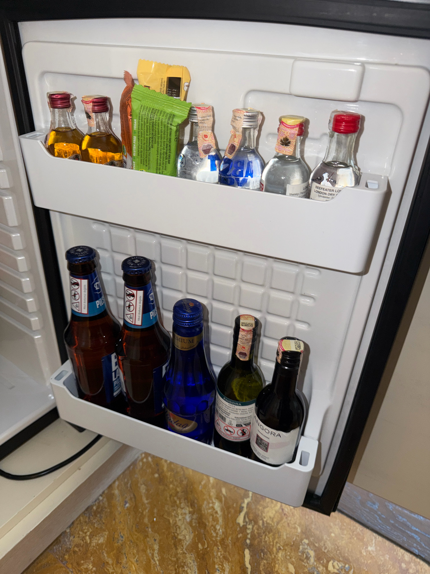An open, white-faced mini-fridge is seen from a slightly elevated, frontal perspective. The interior of the fridge is divided into two shelves. The top shelf, a white plastic tray, contains seven small bottles of alcohol, a green package, and a yellow package. From left to right, the bottles include two mini-bottles of amber liquid, one with a red cap and one with a gold cap, followed by three clear mini-bottles of spirits, two with clear caps and one with a red cap. The yellow package is to the left of the green package, which appears to be a snack bar. The bottom shelf of the fridge, also white plastic, holds six bottles. From left to right, there are two dark brown bottles with blue labels and blue caps, one blue glass bottle with a yellow label and a silver cap, and three dark brown bottles with white labels and silver caps. The fridge door is ajar, revealing the black interior frame and a hint of the exterior. The floor in front of the fridge is covered with a marble-patterned laminate flooring. The scene is set indoors.